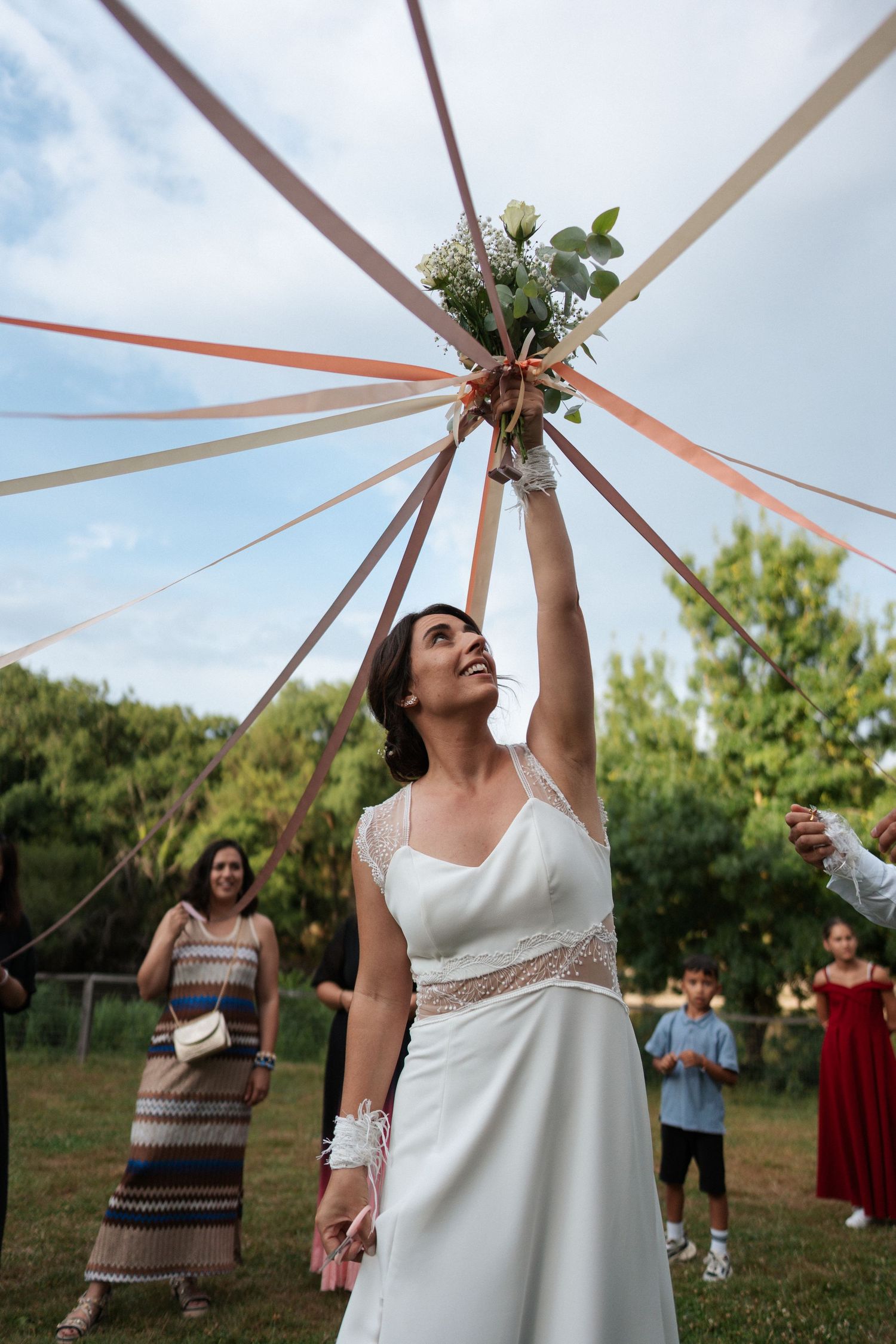 Bride in white gown tossing bouquet with ribbons at outdoor wedding celebration with guests watching.