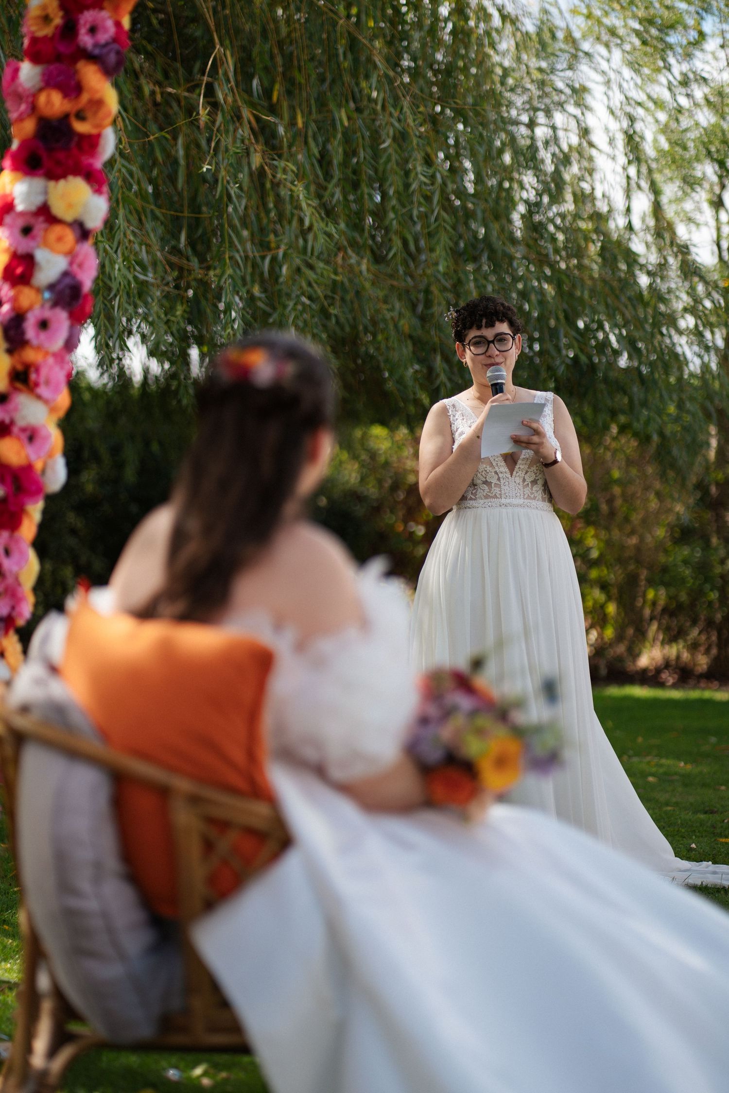 Two brides in white gowns share an outdoor wedding ceremony beside a colorful floral arch and weeping willow tree.