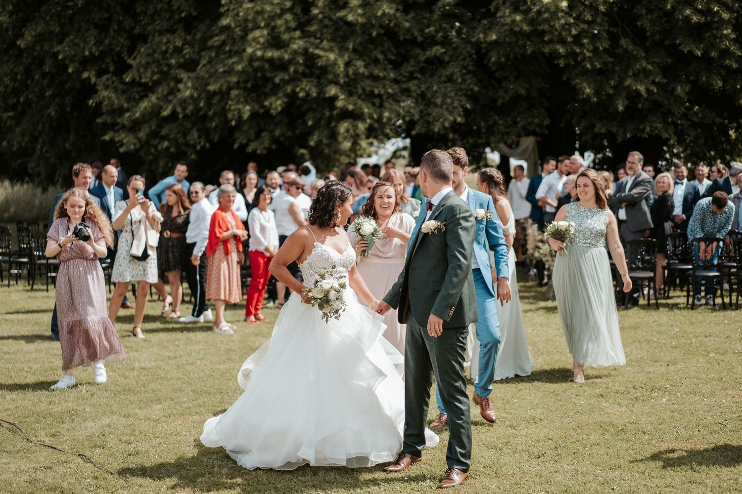 Bride and groom share a romantic moment during outdoor wedding ceremony with guests celebrating on sunny green lawn.