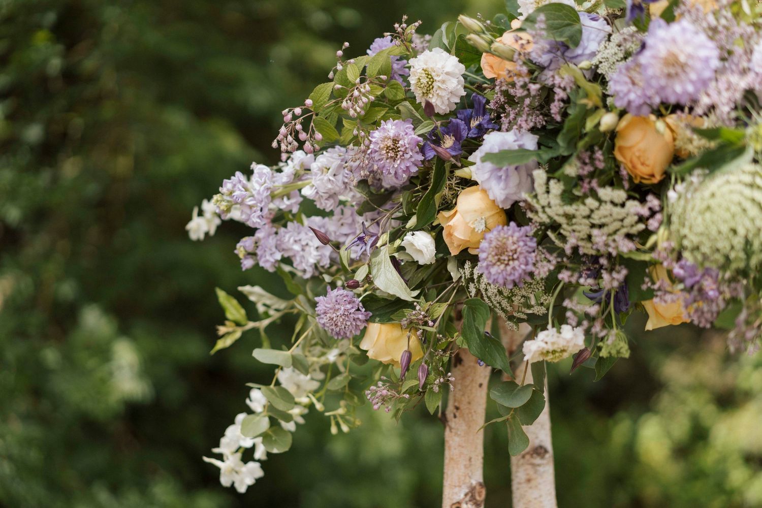 Composition florale pour arche de cérémonie laïque – Bouquet de fleurs mauves et jaunes fixé sur structure en bois, style mariage champêtre.