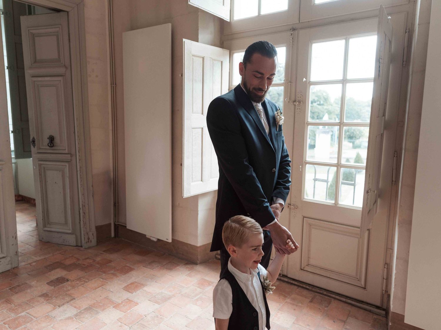 Un homme élégamment vêtu tient la main d'un jeune garçon dans un couloir ensoleillé d'un château.