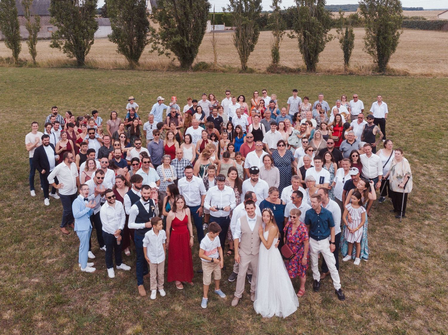 Photo de groupe de mariage vue par drone – Mariés et invités alignés en extérieur dans un champ, formation en cœur pour un cliché original.