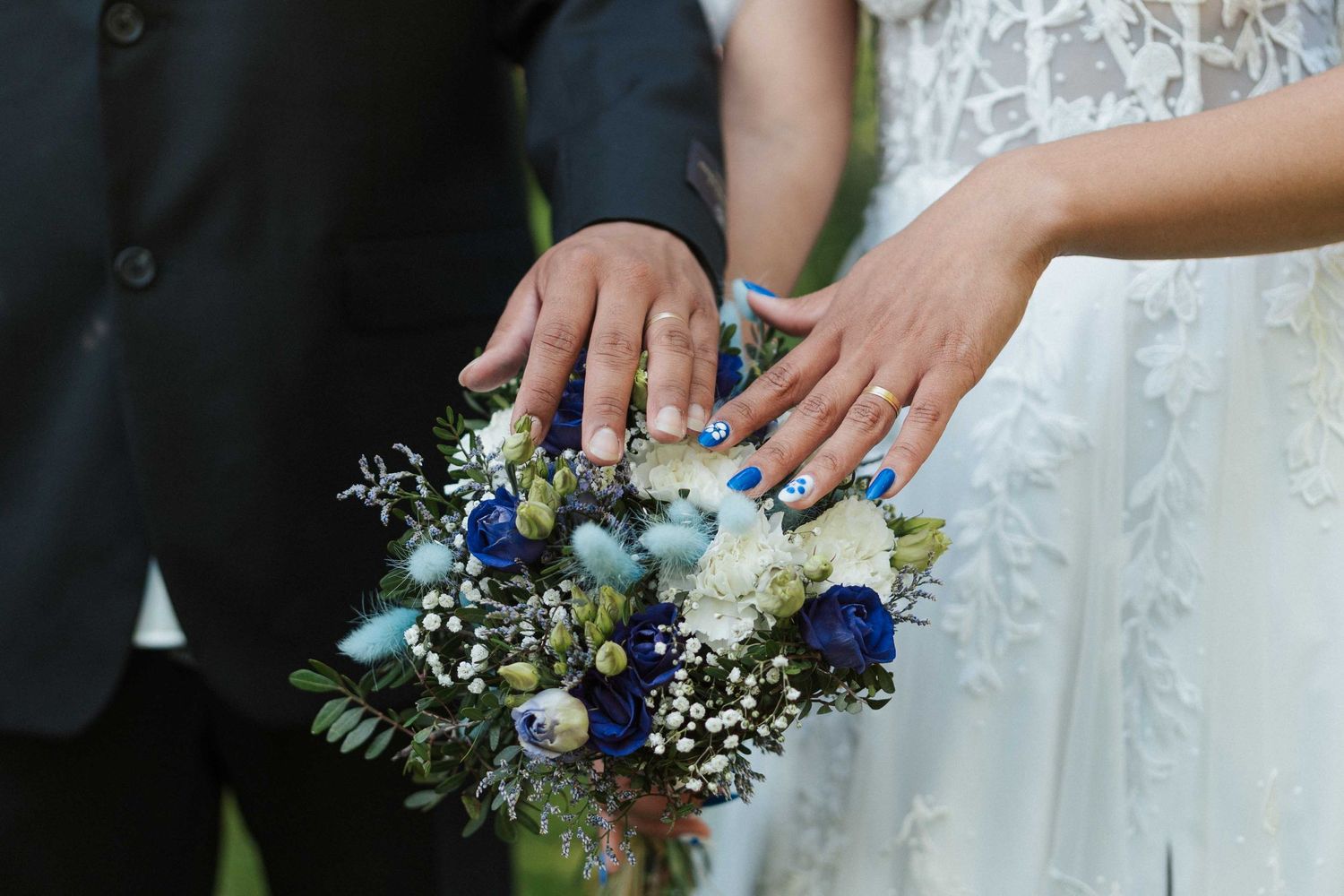 Des jeunes mariés exhibent leurs alliances sur un bouquet de fleurs bleues et de roses blanches.