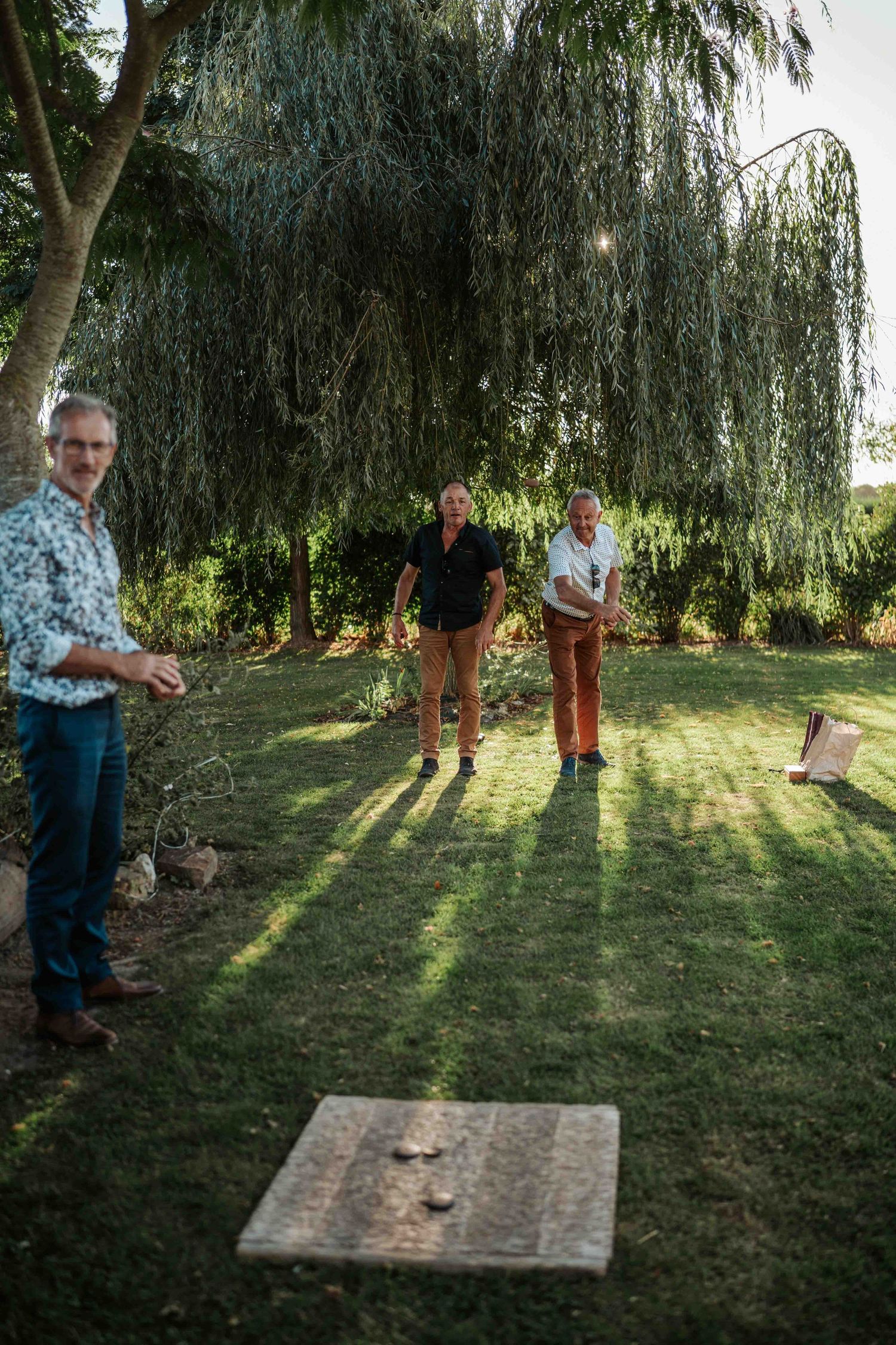 Trois invités jouent à des jeux de plein air sous un saule pleureur lors d'une réception de mariage en extérieur au crépuscule.