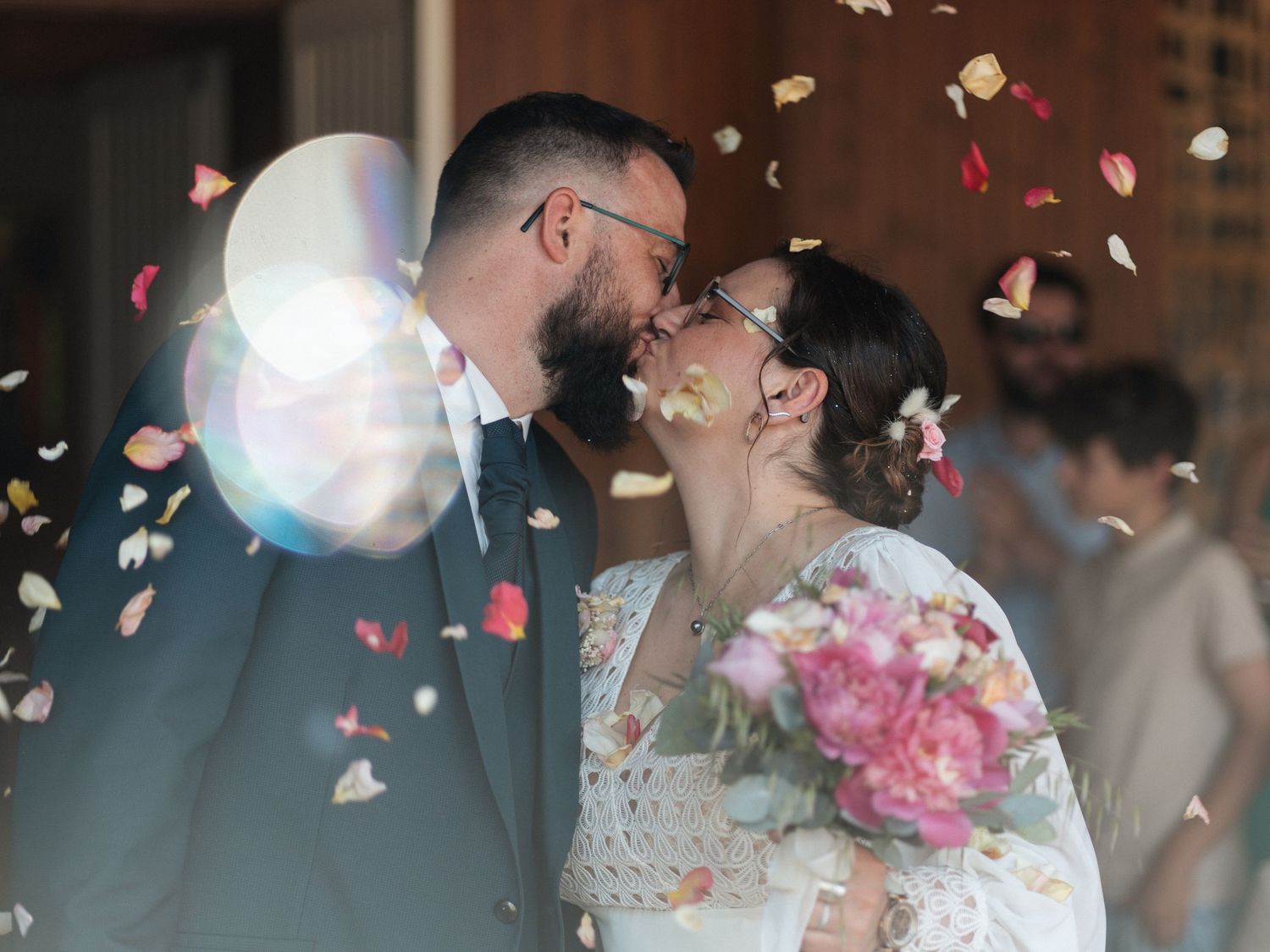 Newlyweds share a kiss surrounded by colorful flower petal confetti, bride holding pink bouquet.