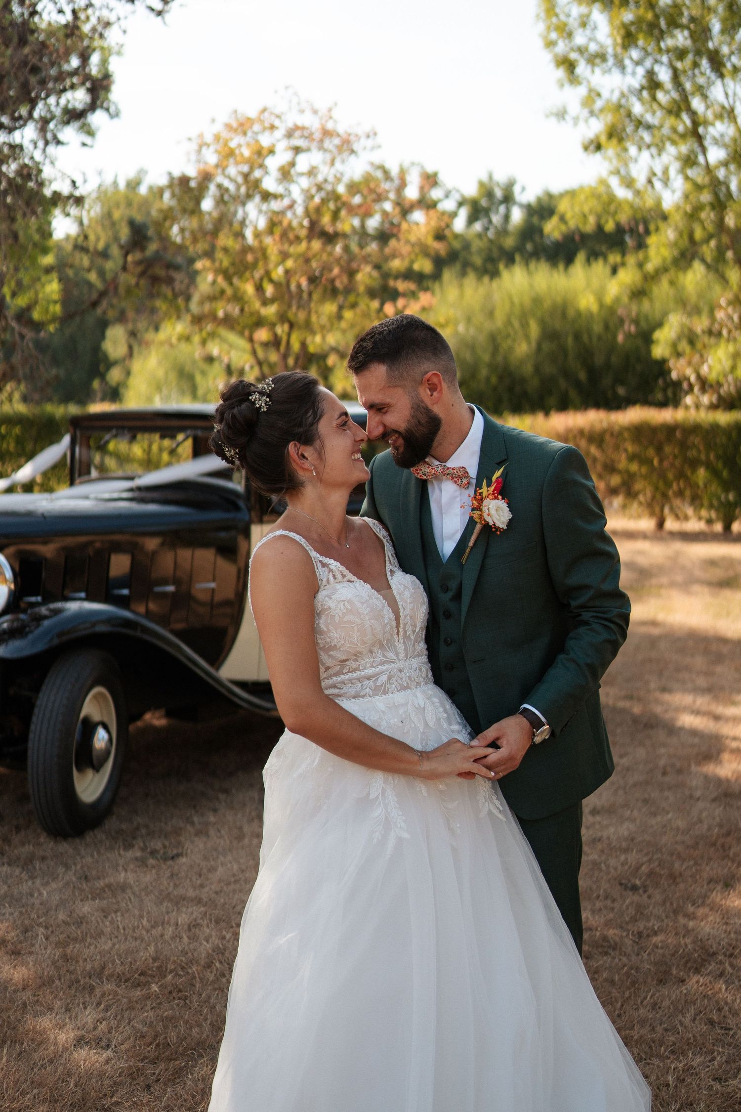 Bride and groom share a romantic moment in front of a vintage black car, surrounded by lush greenery on their wedding day.