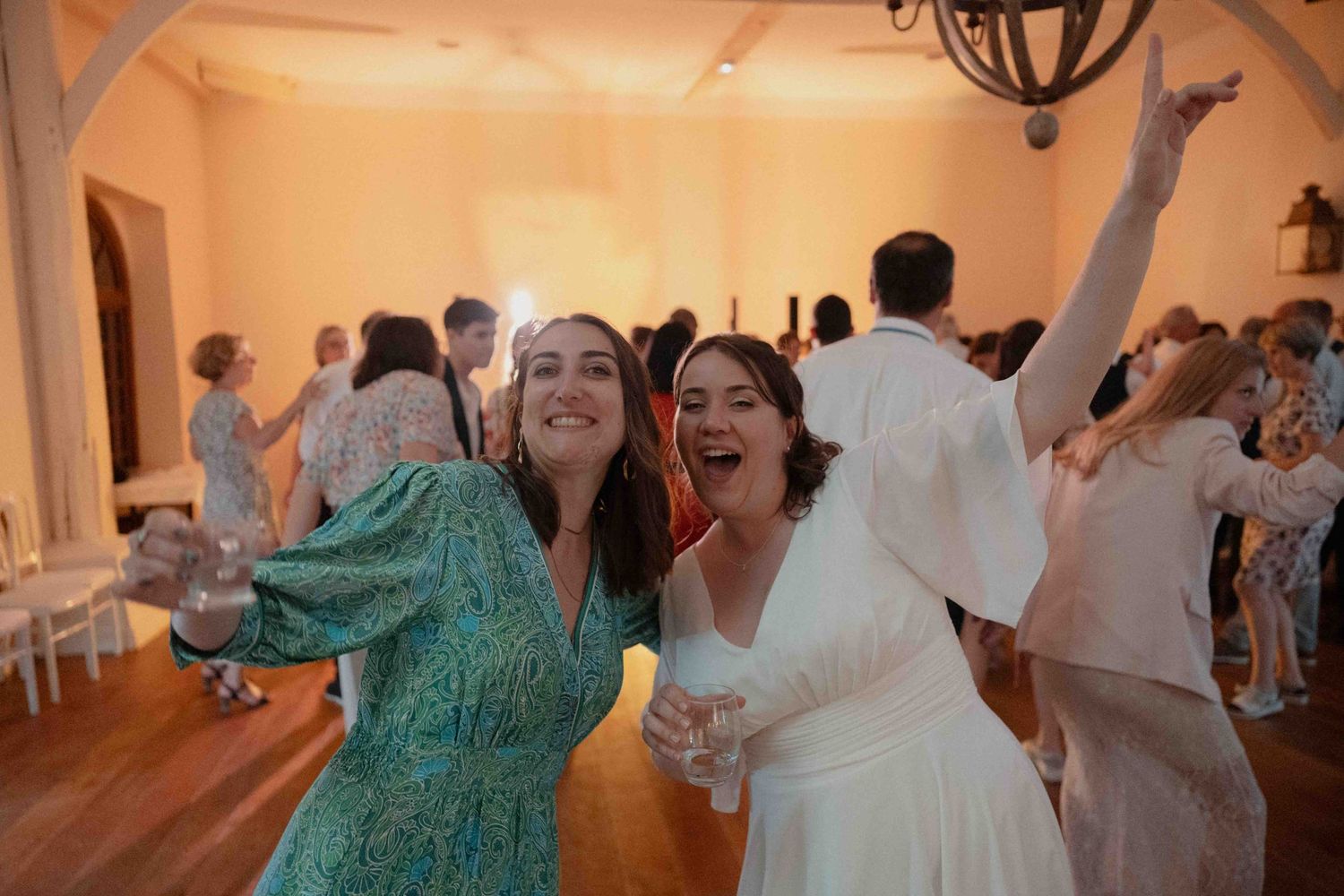 Deux femmes sourient et dansent joyeusement lors d'une réception de mariage en intérieur, baignée d'une lumière chaleureuse.
