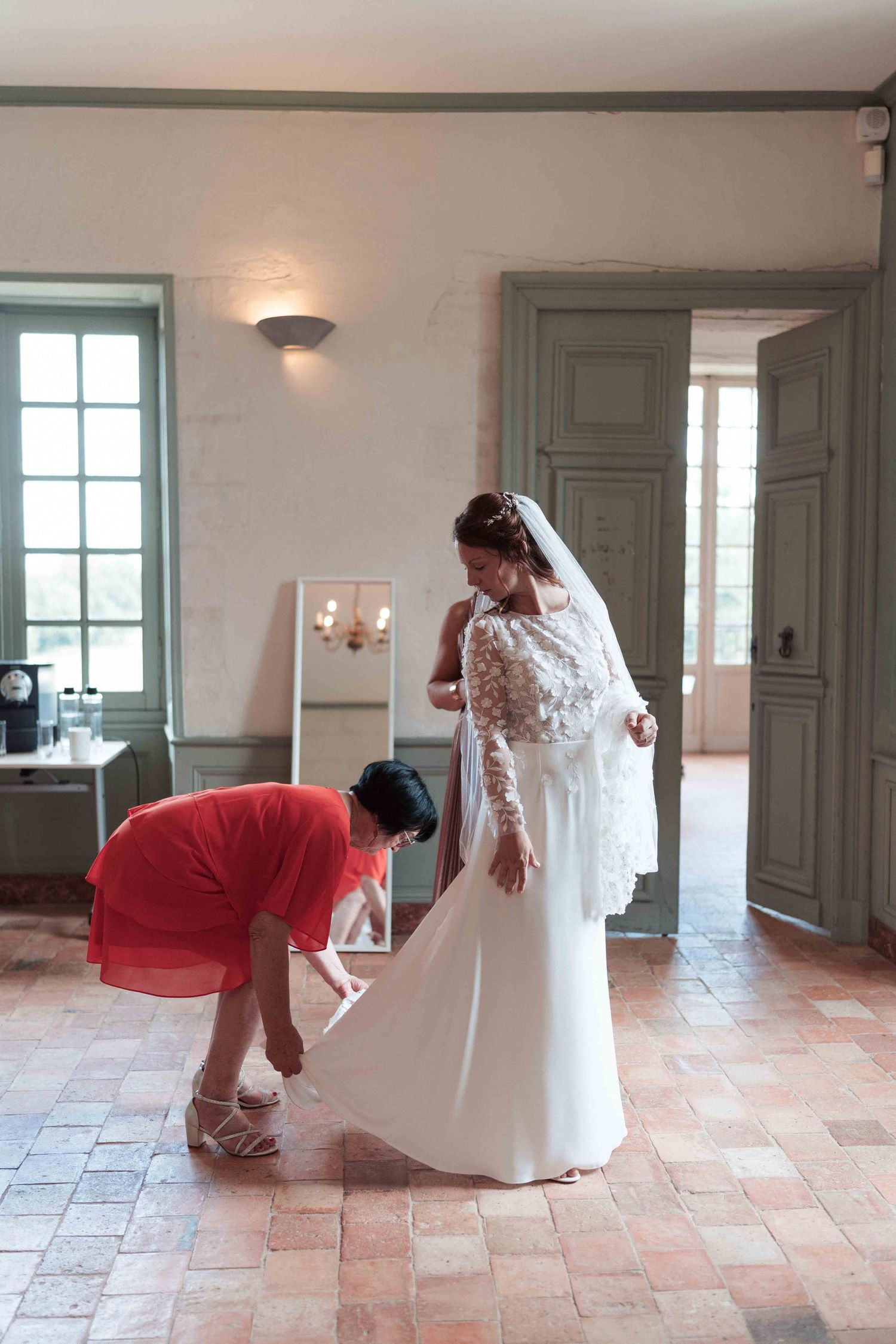 Une demoiselle d'honneur en robe rouge aide la mariée à se chausser dans une élégante suite nuptiale d'un château français.