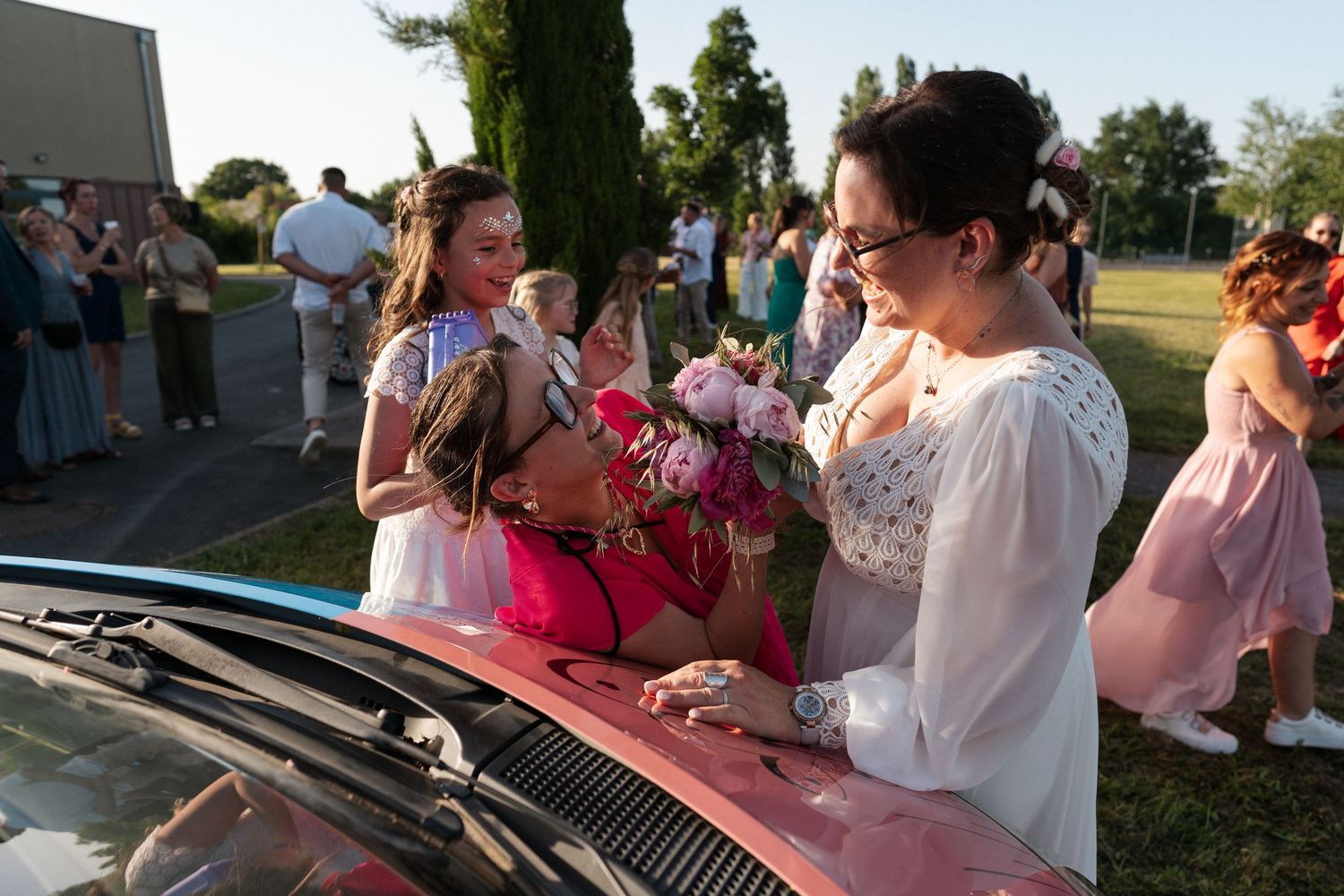Women in elegant dresses gather around a red car at an outdoor wedding celebration, exchanging flowers and greetings.