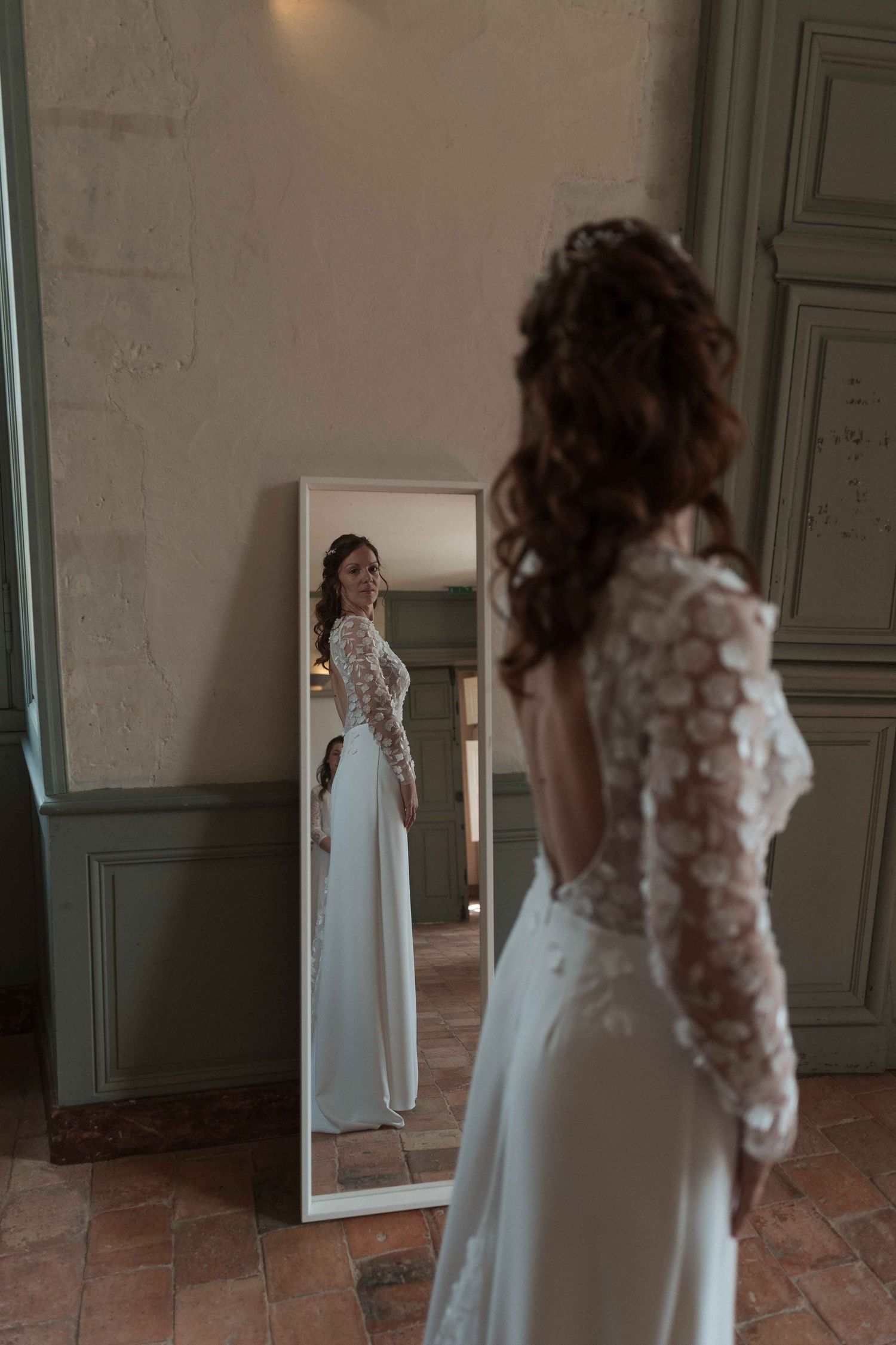 La mariée, vêtue d'une robe de mariée à dos en dentelle, admire son reflet dans un miroir en pied.