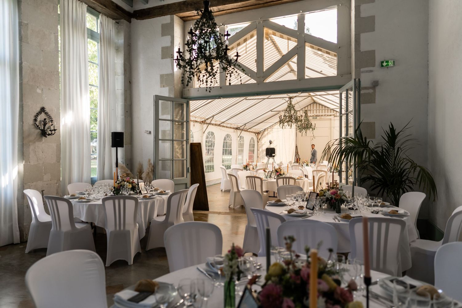 Salle de réception lumineuse de l’Orangerie de Maulny, décorée pour un mariage avec nappes blanches et lustres élégants.