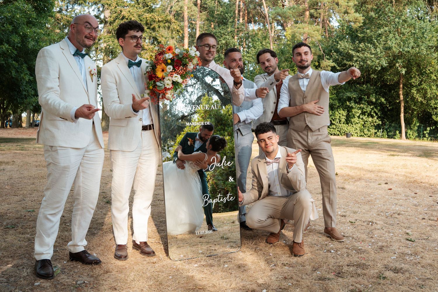 Wedding party group photo with groom and groomsmen in tan suits holding a decorative sign outdoors.