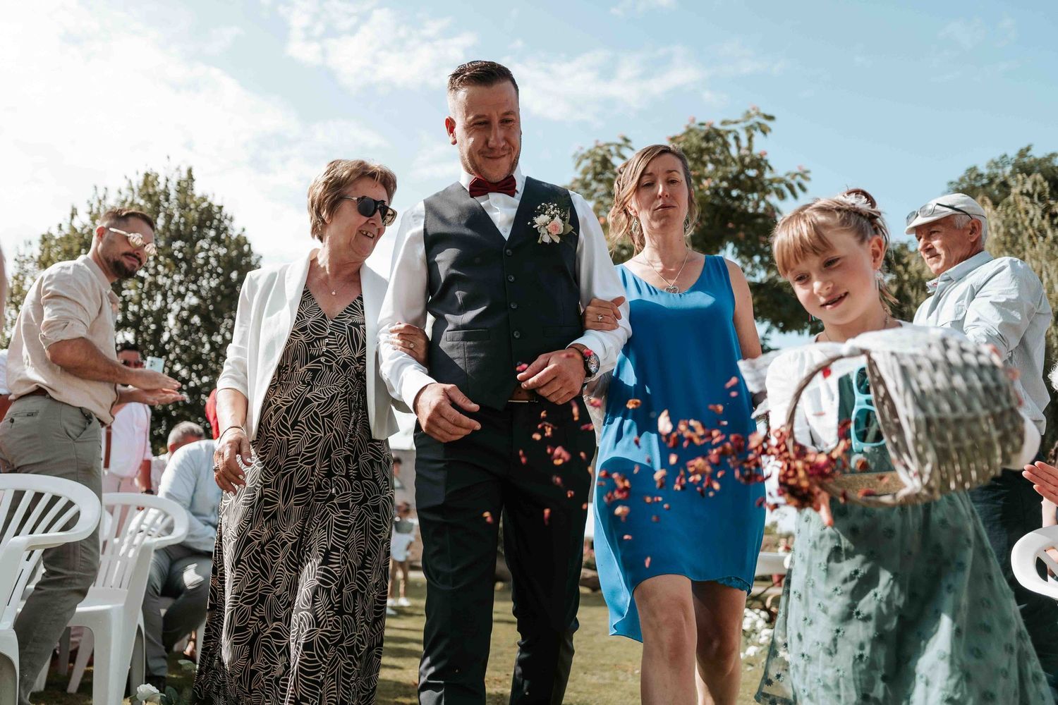Le marié descend l'allée de mariage en plein air, accompagné de sa famille, tandis que la petite fille d'honneur jette des pétales