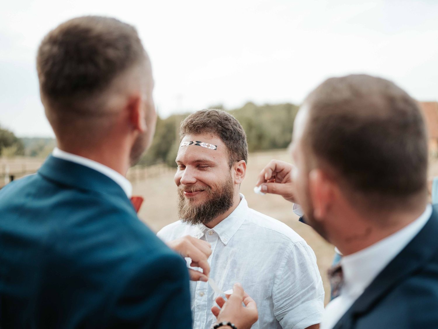 Un témoin souriant et barbu, le visage couvert de confettis, discute avec les invités lors d'un mariage en plein air.