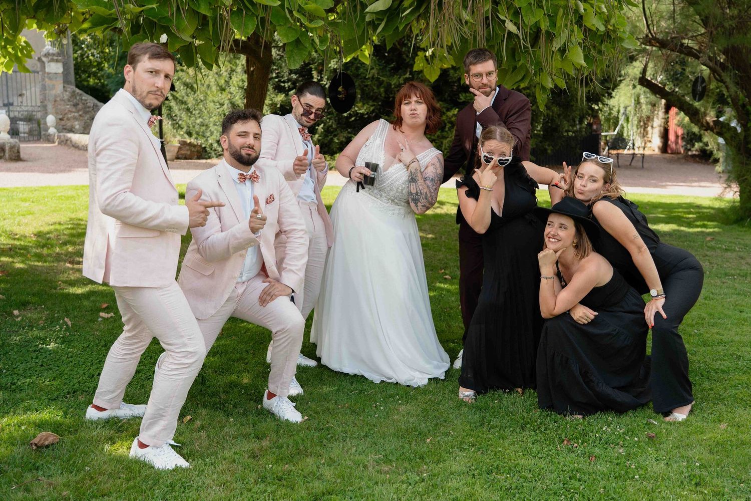 Le cortège nuptial pose de manière ludique en extérieur sur l'herbe verte, la mariée en robe blanche et les invités en tenue de soirée.