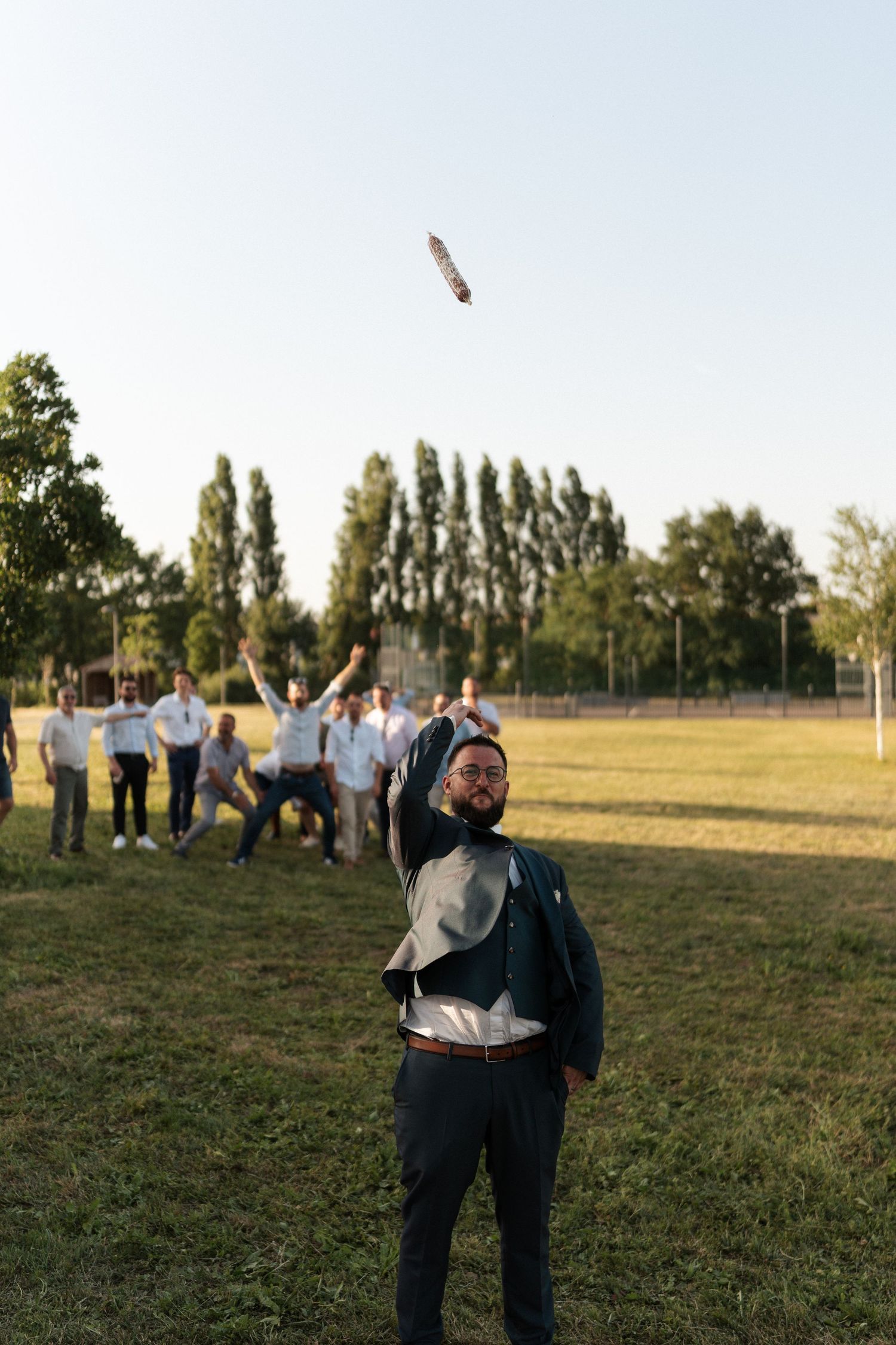 Groom tosses bouquet to groomsmen gathered on green field during outdoor wedding celebration on sunny day.