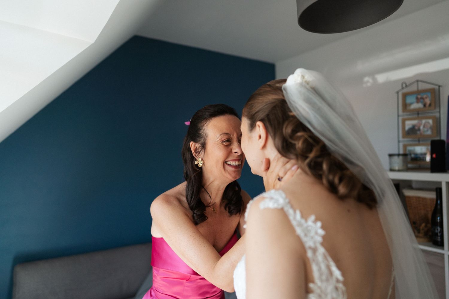 A bride in a white lace gown and veil shares a joyful moment with a woman in a pink dress before the wedding.