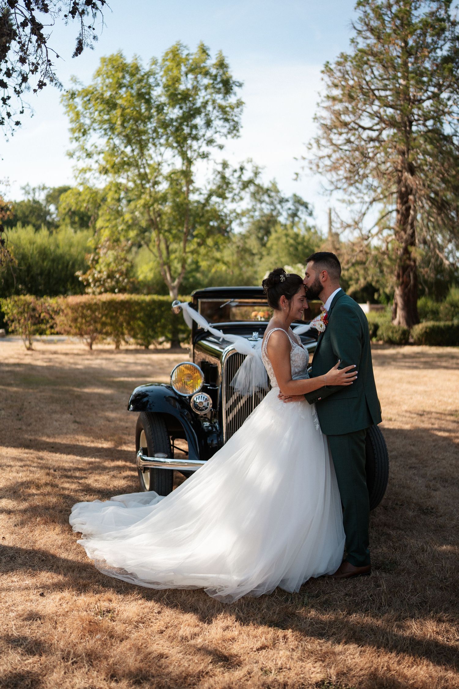 Bride and groom share a romantic moment leaning against a vintage classic car surrounded by trees on their wedding day.