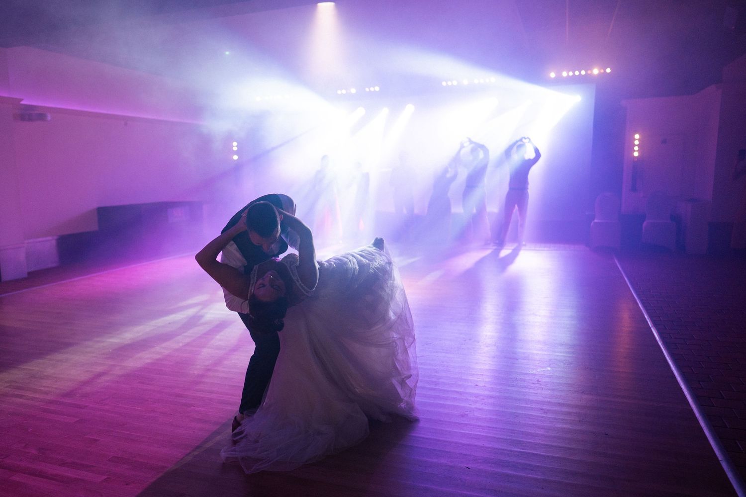 Bride and groom share a romantic dip kiss on a foggy dance floor with dramatic purple and blue stage lighting.