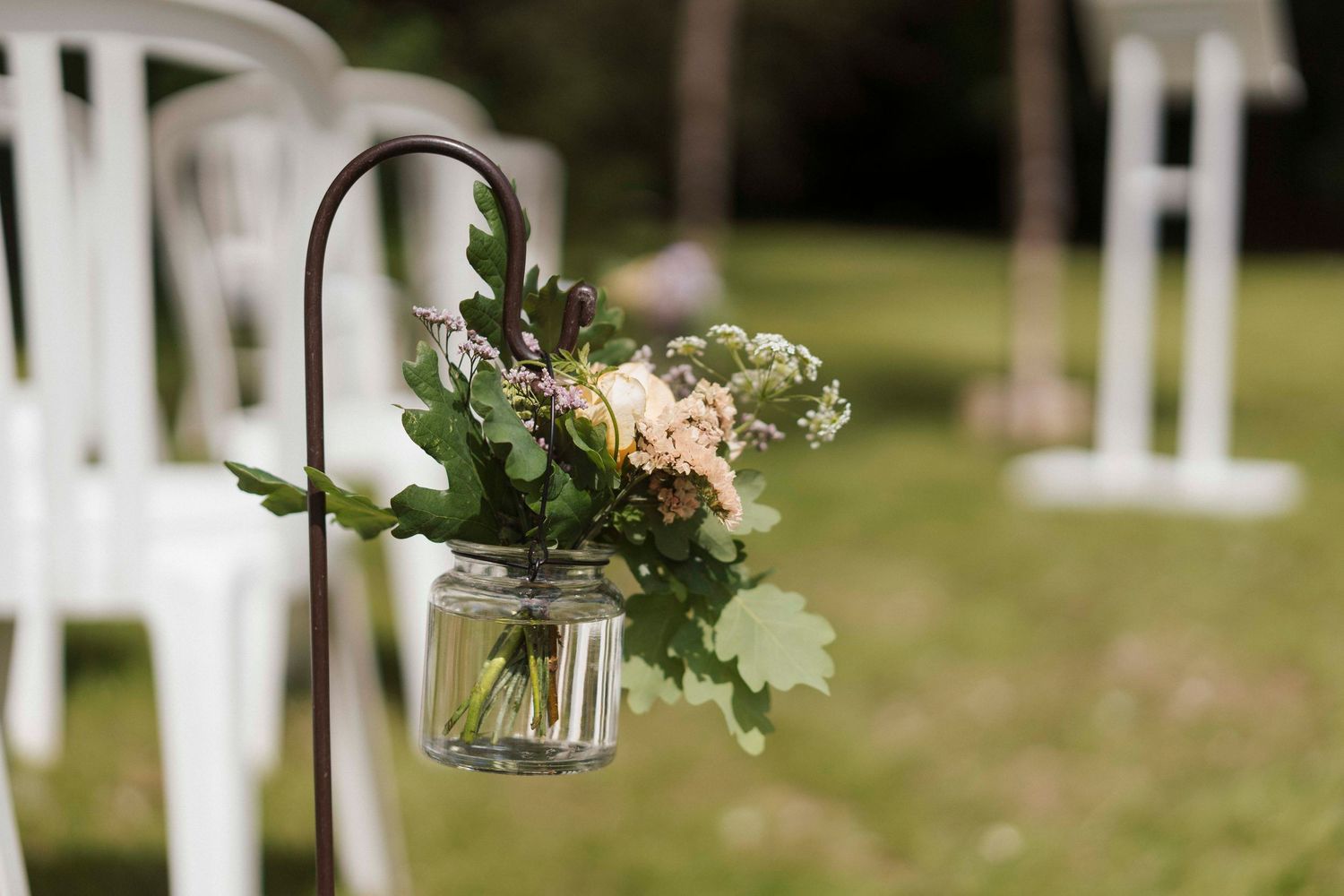 Petit bouquet suspendu dans un bocal en verre – Décoration d’allée de cérémonie laïque en extérieur, fleurs pastel et feuillage vert.
