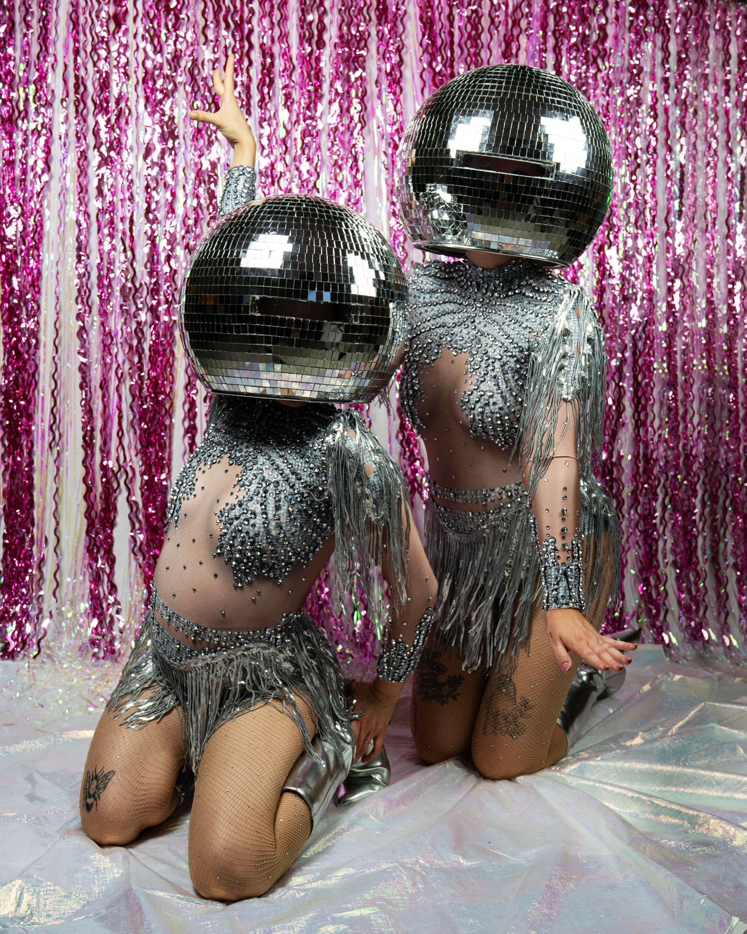Two performers kneel in sparkly silver fringe costumes with large disco ball helmets against pink tinsel backdrop.