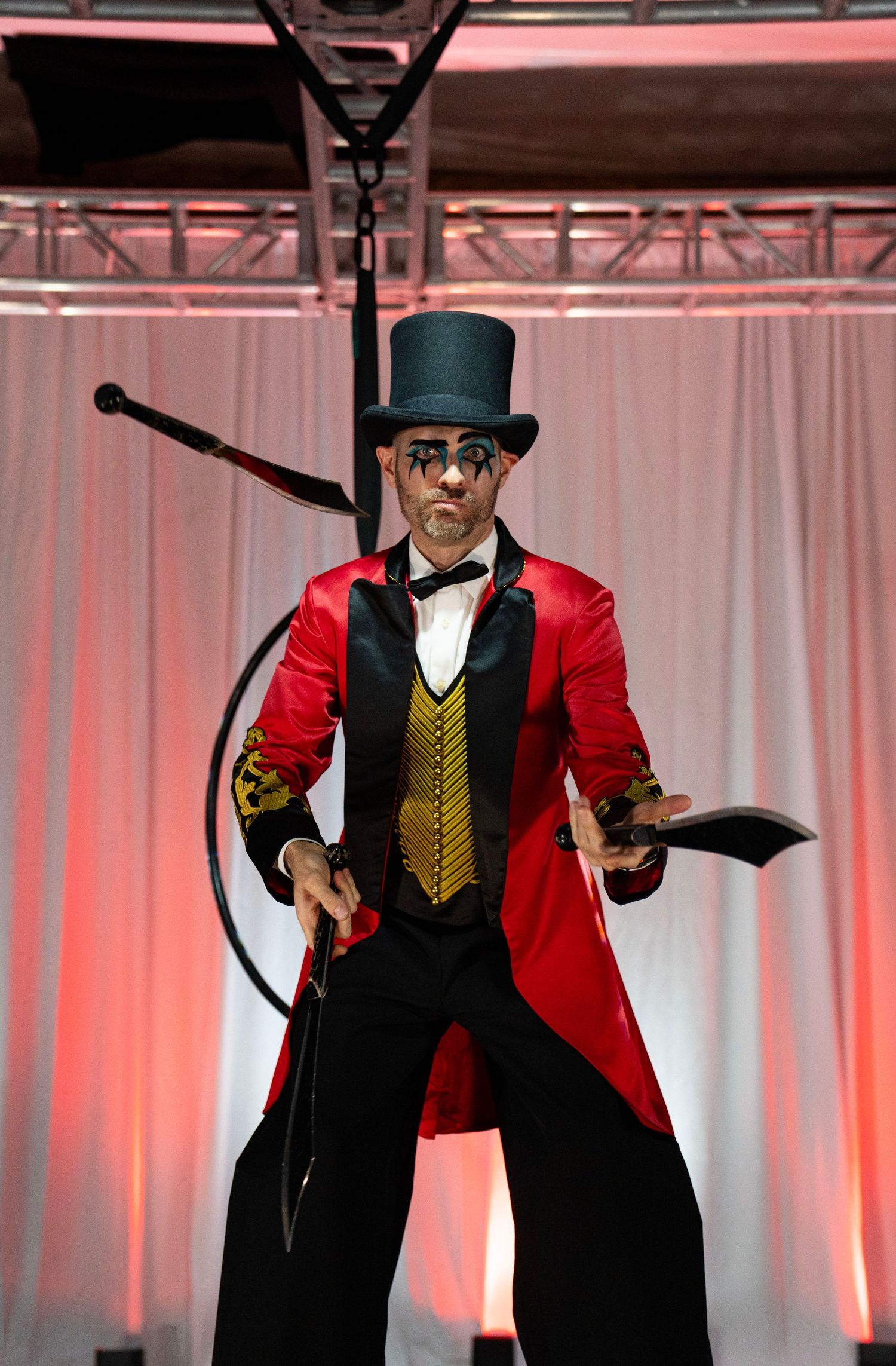 Male circus ringmaster in red tailcoat juggles knives while performing on stilts on a lit stage.