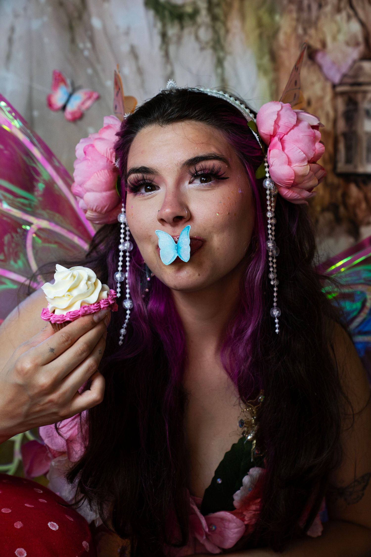Woman dressed as fairy with pink peony hair, iridescent wings holds cupcake with blue butterfly on lips. Fantasy Photoshoot