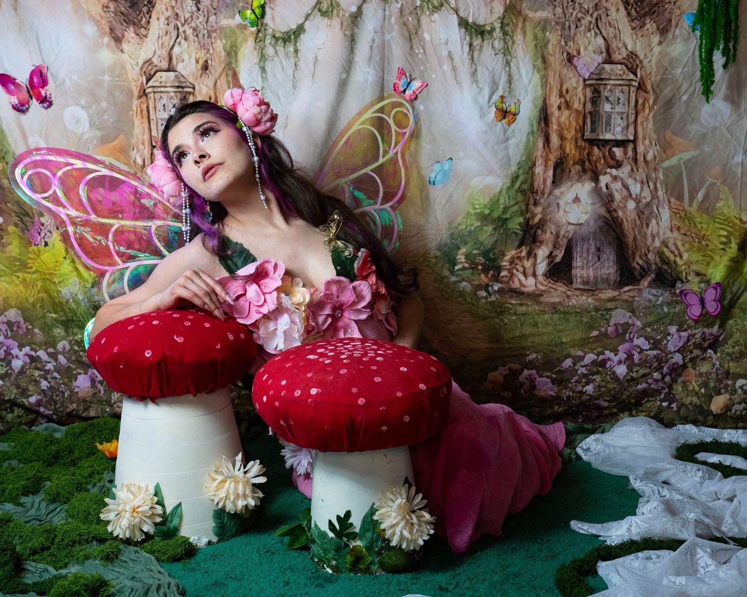 Fairy costume model with pink butterfly wings reclines on red toadstools in whimsical enchanted forest scene. Fantasy Photoshoot