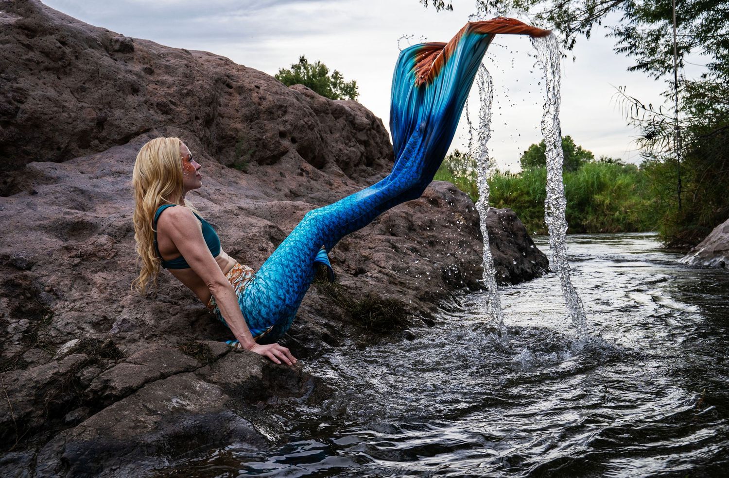 A woman in a blue mermaid tail lifts her fin as water cascades off it onto the rocks. Fantasy Photoshoot