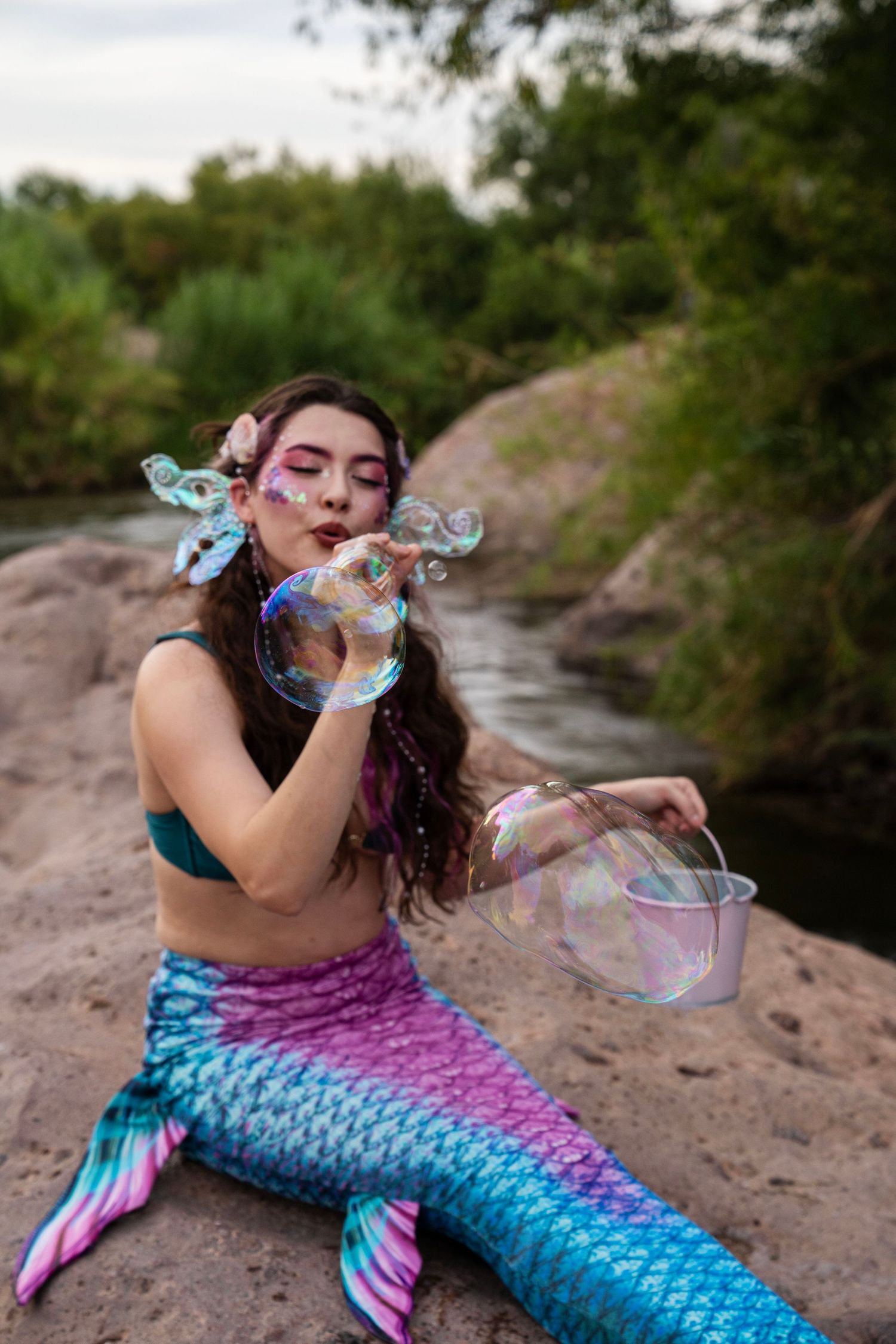 Model in mermaid costume with iridescent tail blowing large soap bubbles while sitting on river rocks. Fantasy Photoshoot