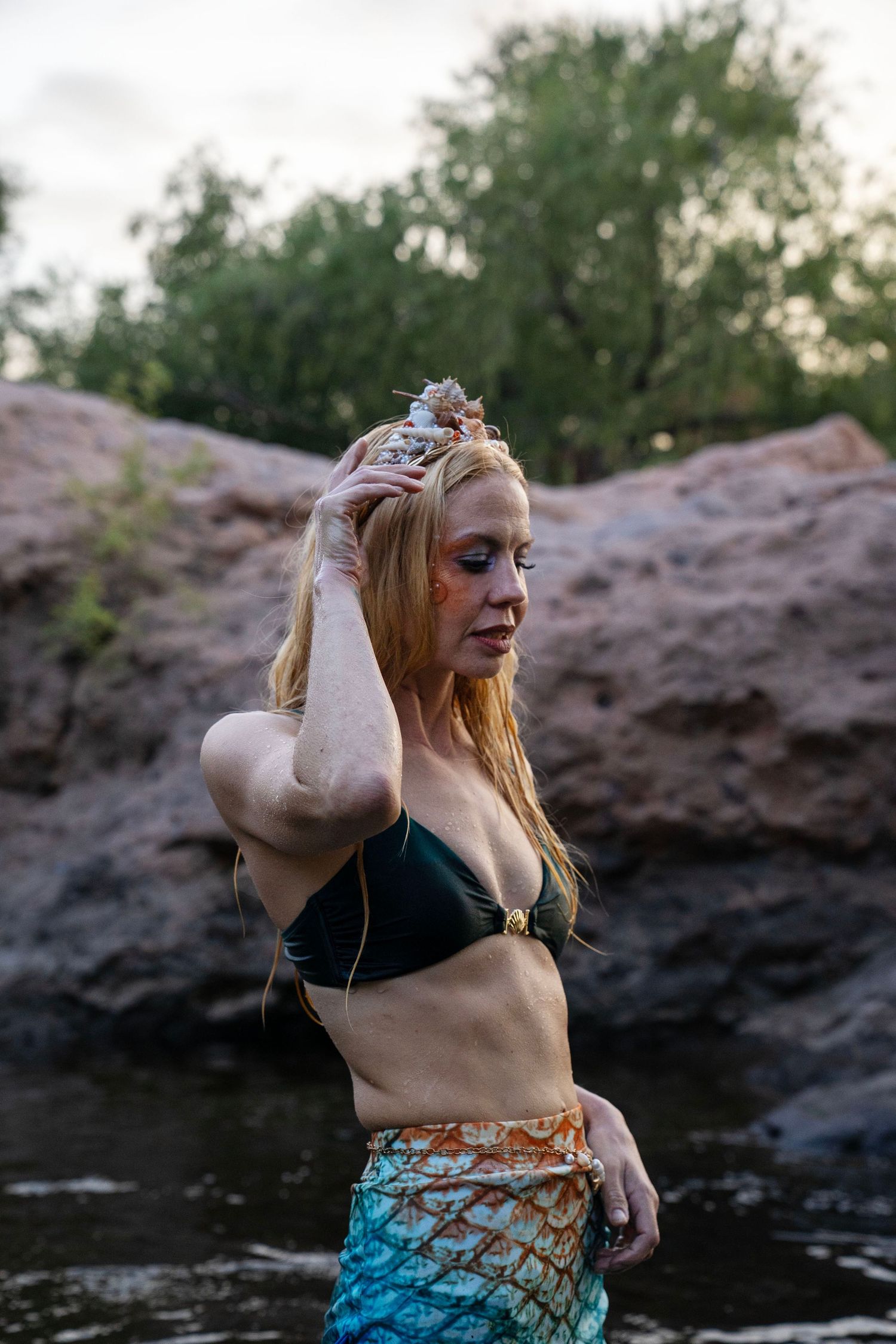 A blonde woman in a mermaid costume with colorful tail and dark bikini top poses near rocky riverbank at dusk. Fantasy Photoshoot