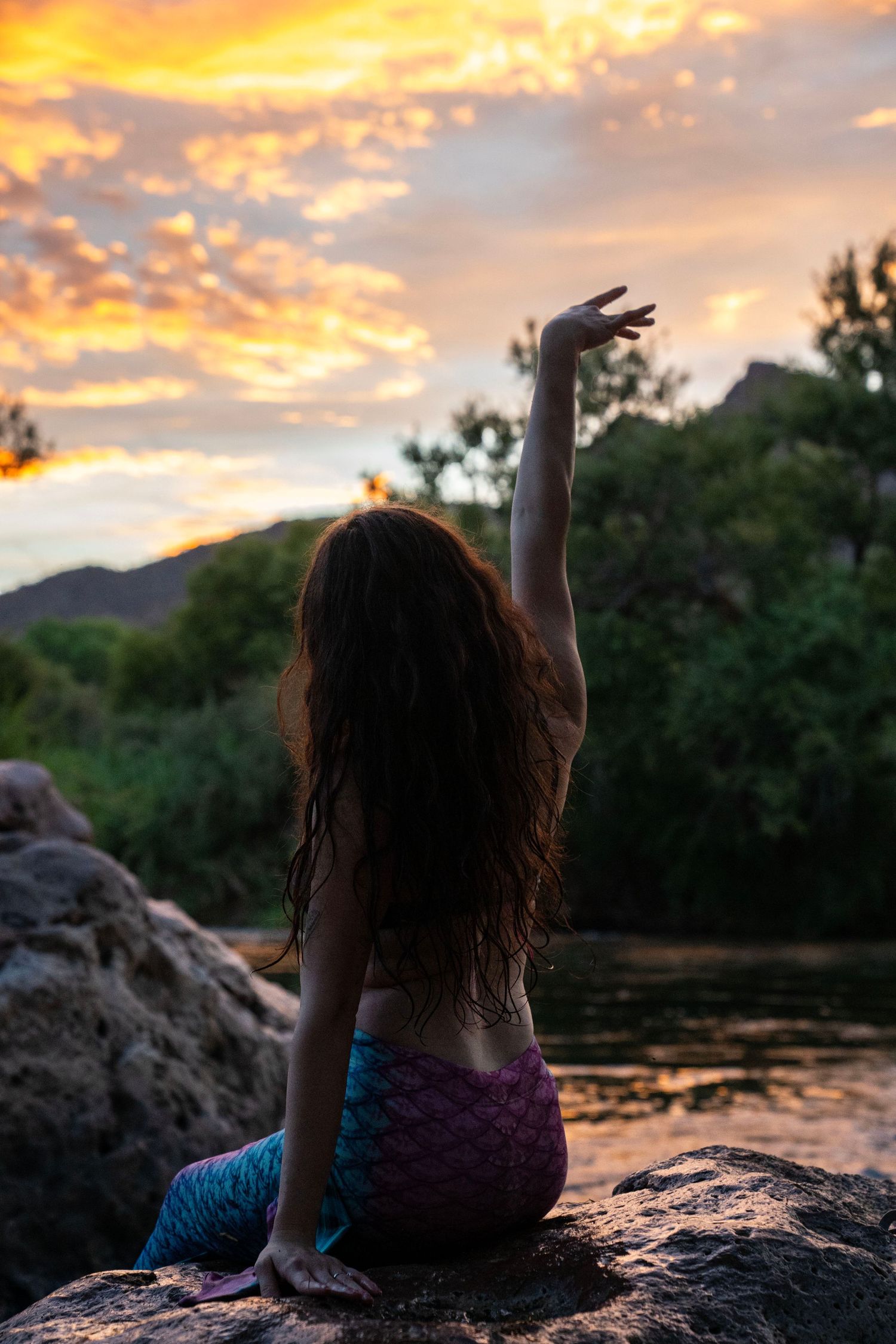 A woman in a mermaid costume raises her hand toward a stunning orange and pink sunset sky. Mermaid photoshoot, fantasy photoshoot.