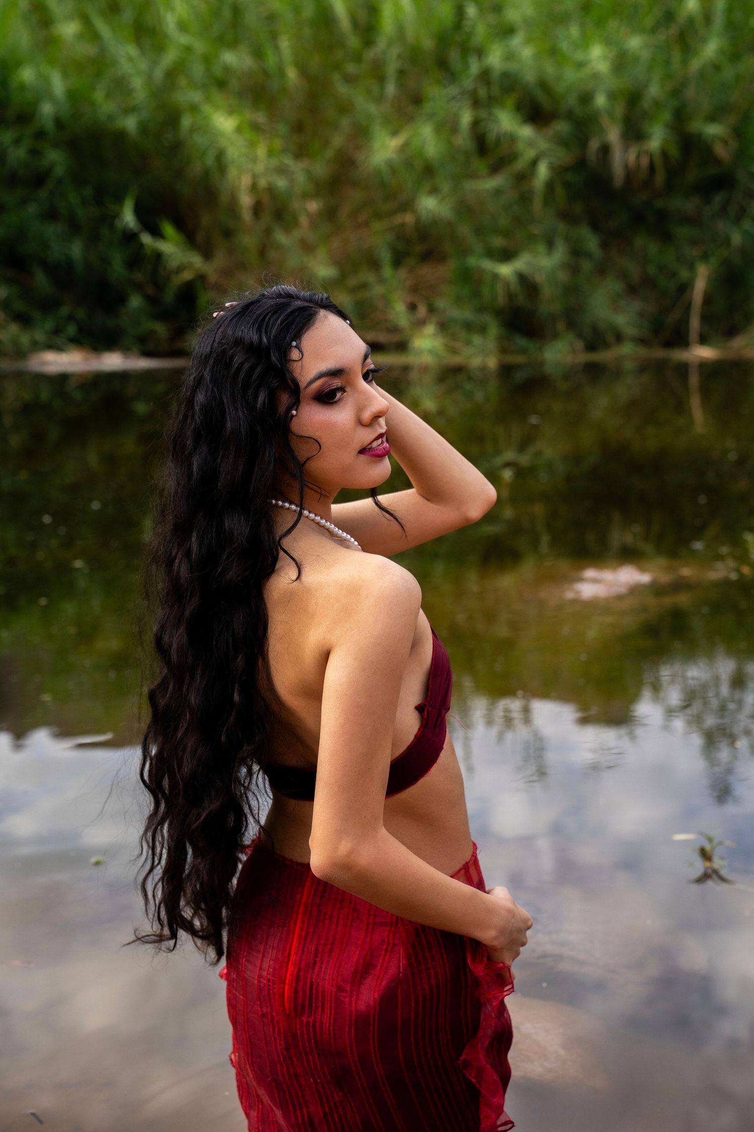 A woman with long black wavy hair poses in a red saree while standing in a shallow river with green foliage behind her.
