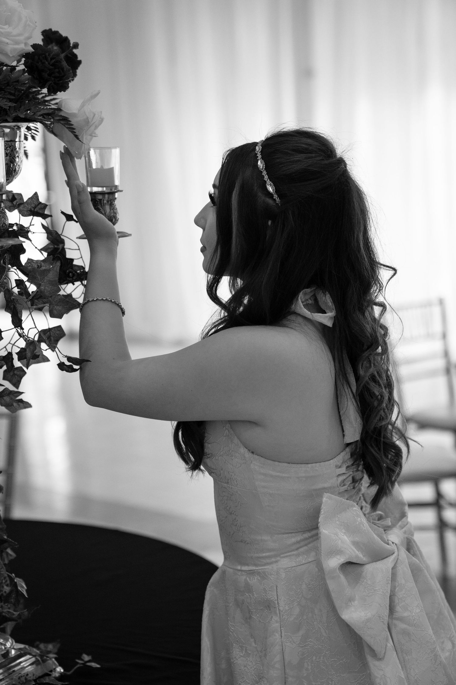 Black and white photo of a bride in a strapless gown with bow, arranging floral centerpiece at wedding reception.