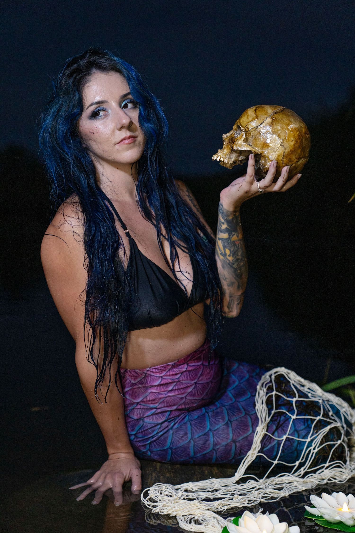 Tattooed woman in mermaid costume holding a skull beside a fishing net and water lilies at night.