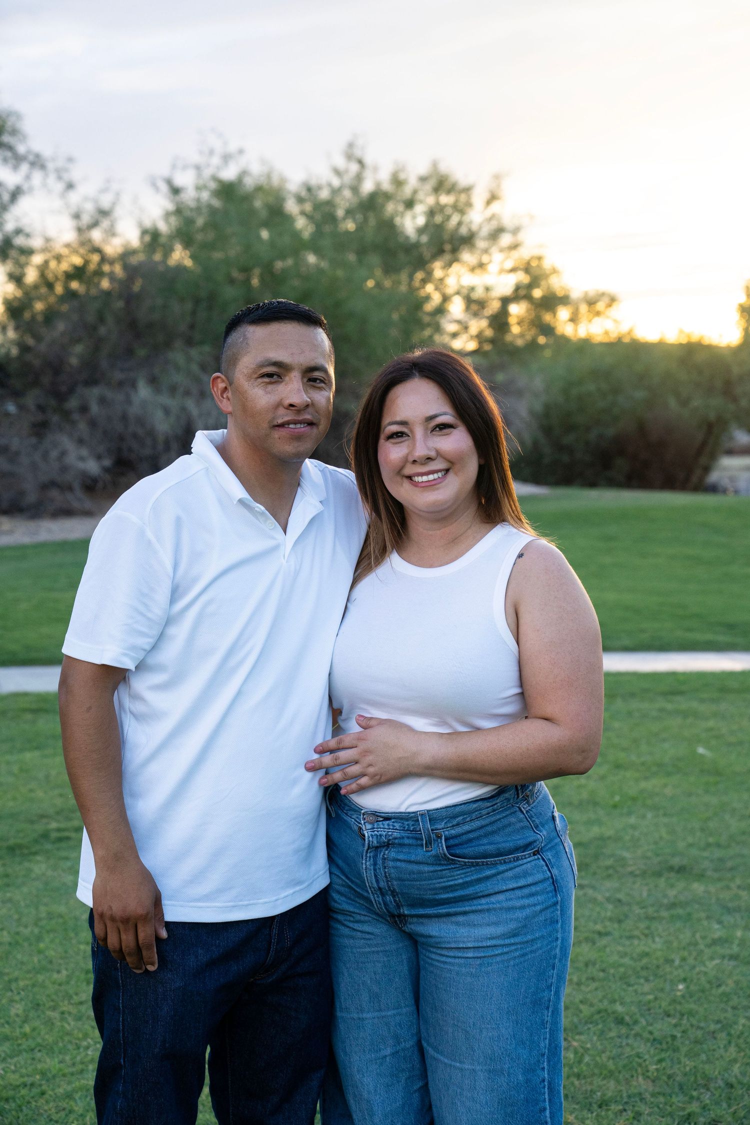 Smiling couple posing together on a green lawn at sunset, wearing white tops and jeans.