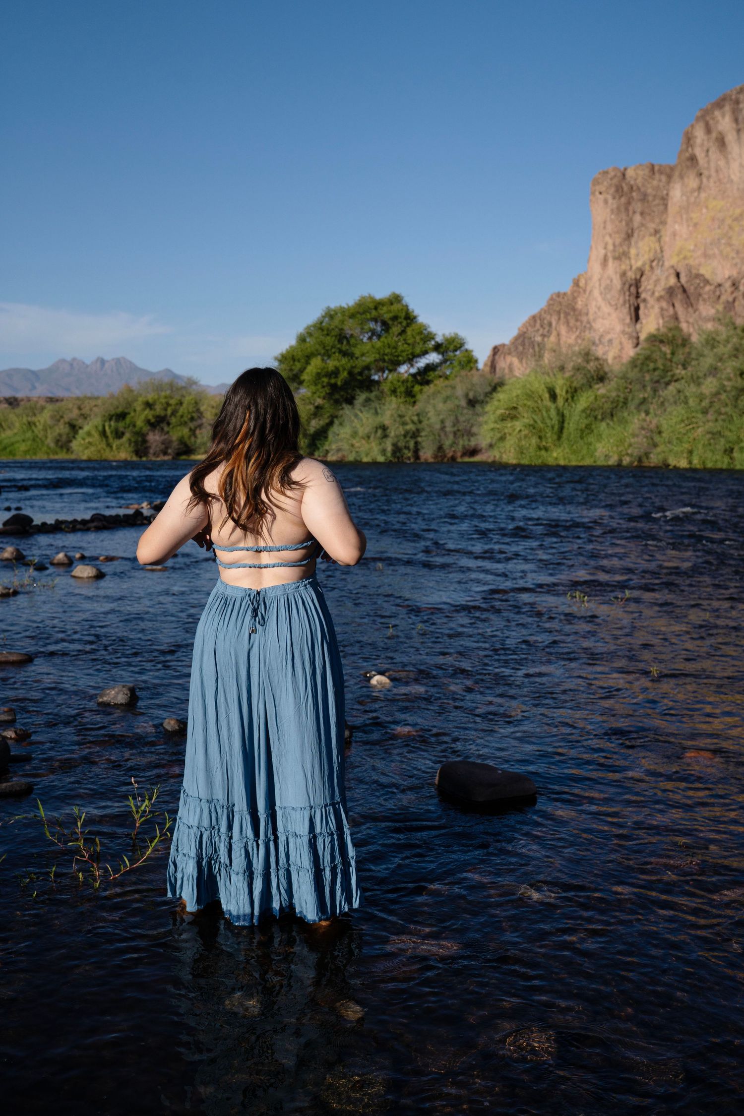 Woman in blue maxi dress standing in a rocky river with desert cliffs and green trees under a clear blue sky.