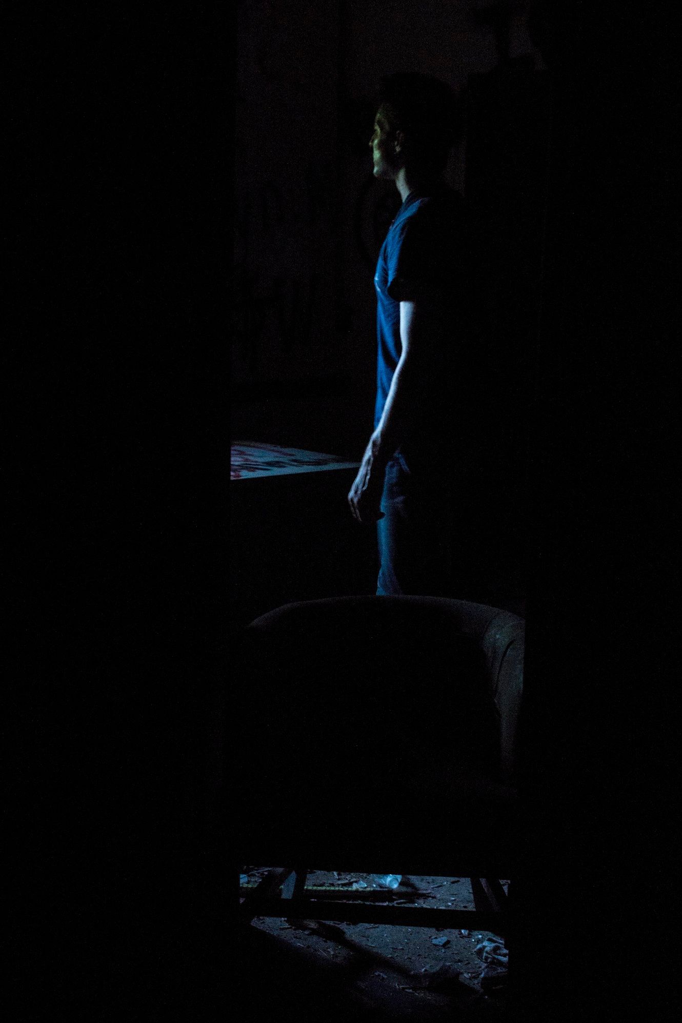 Silhouette of a man in a dark blue t-shirt standing in a dimly lit abandoned room beside an old armchair, lit by faint blue light.