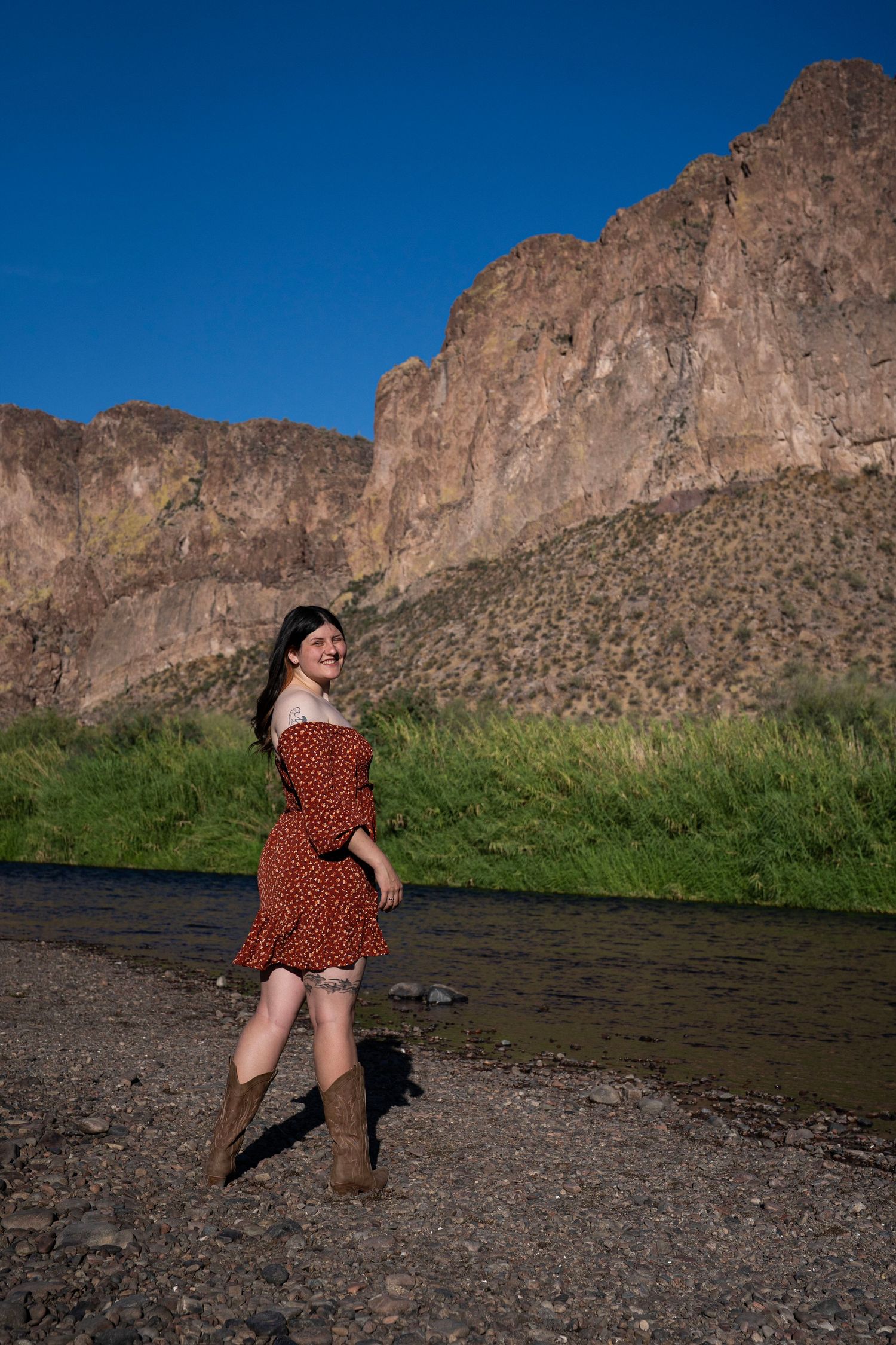 Young woman in red floral mini dress and brown cowboy boots walking along a scenic desert river bank.