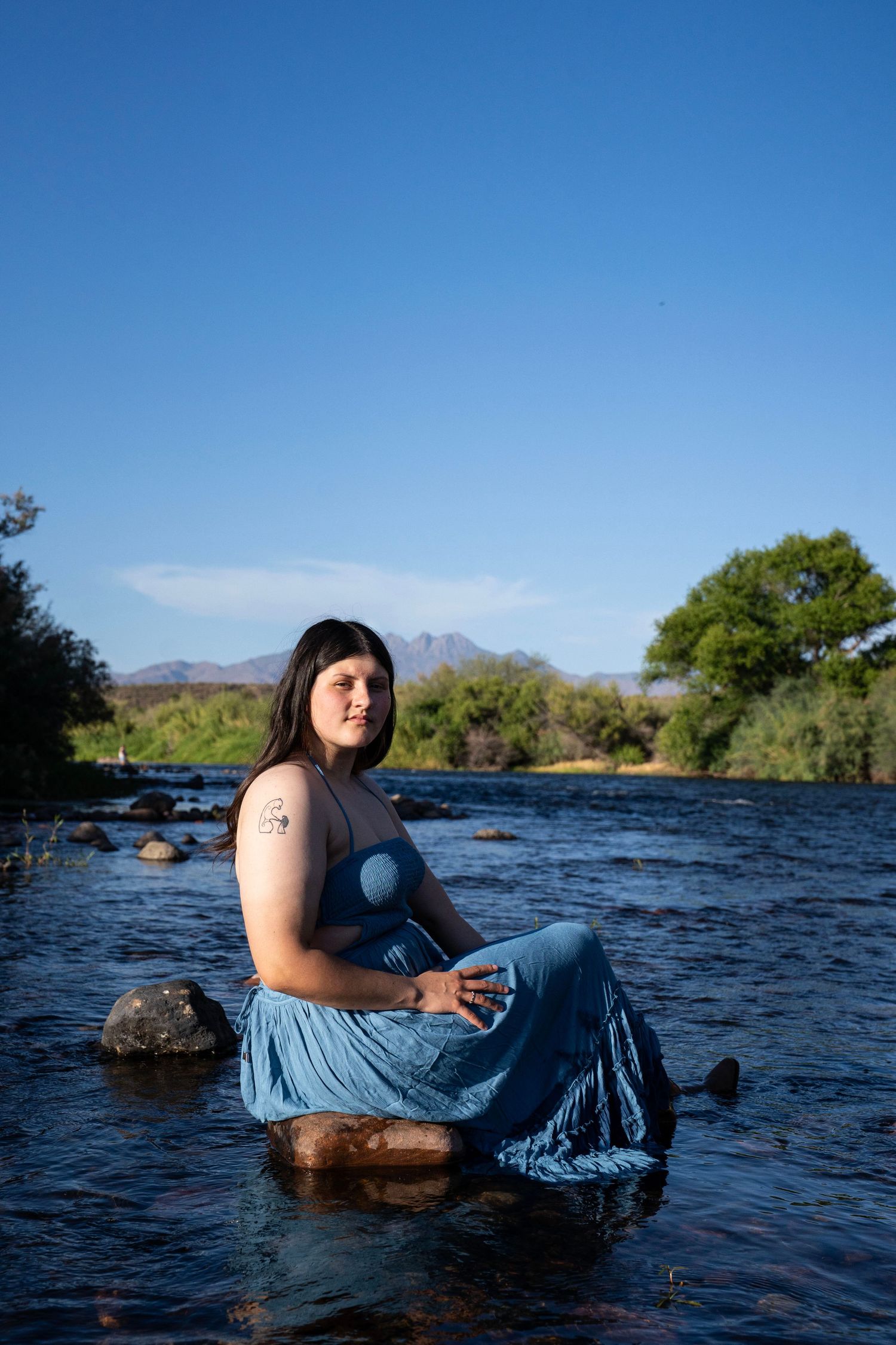 Woman in blue dress sitting on rocks in a scenic river with mountains and trees under a clear blue sky.
