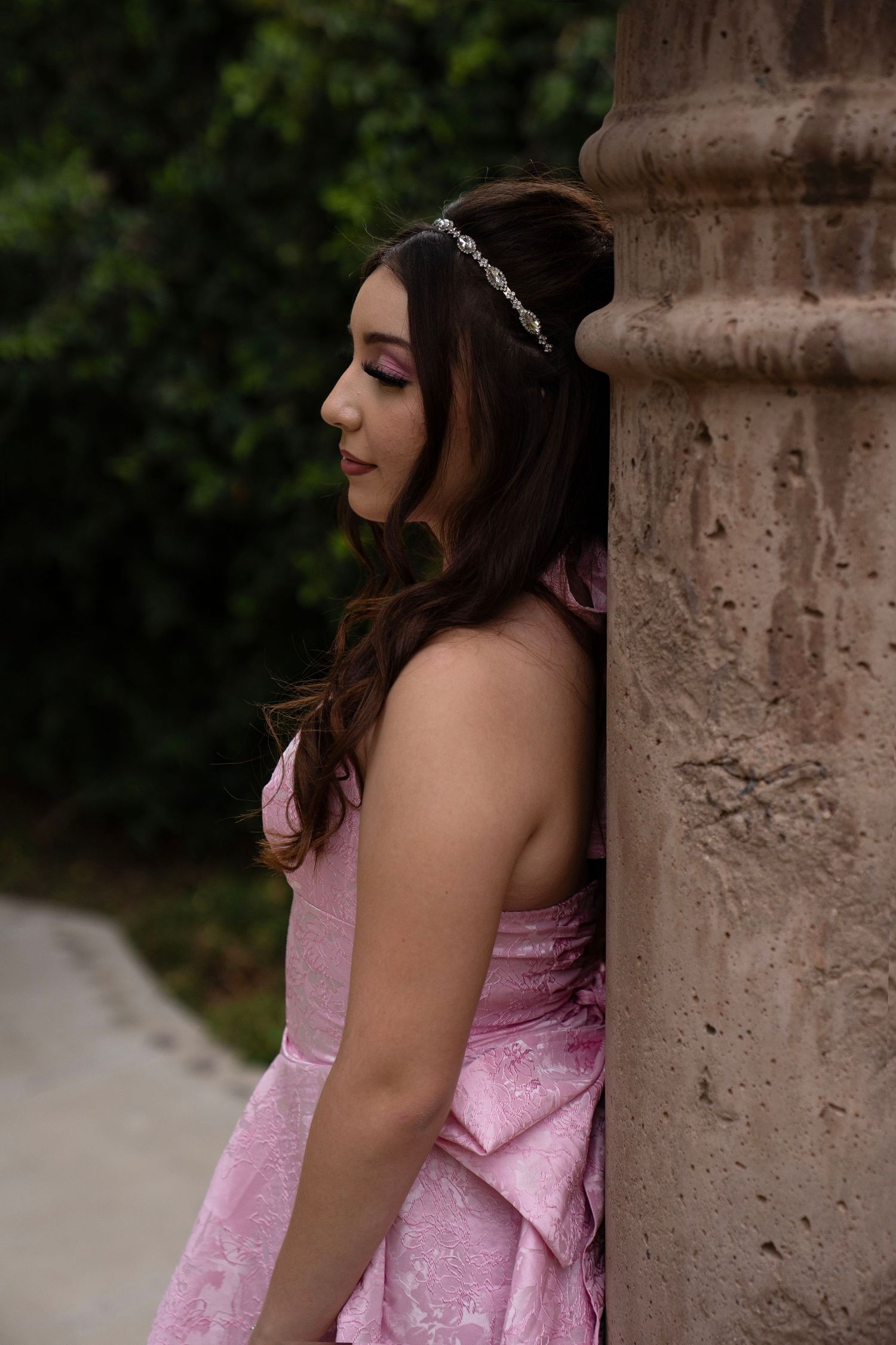 Profile view of brunette woman in pink strapless dress with crystal hair accessory by column.
