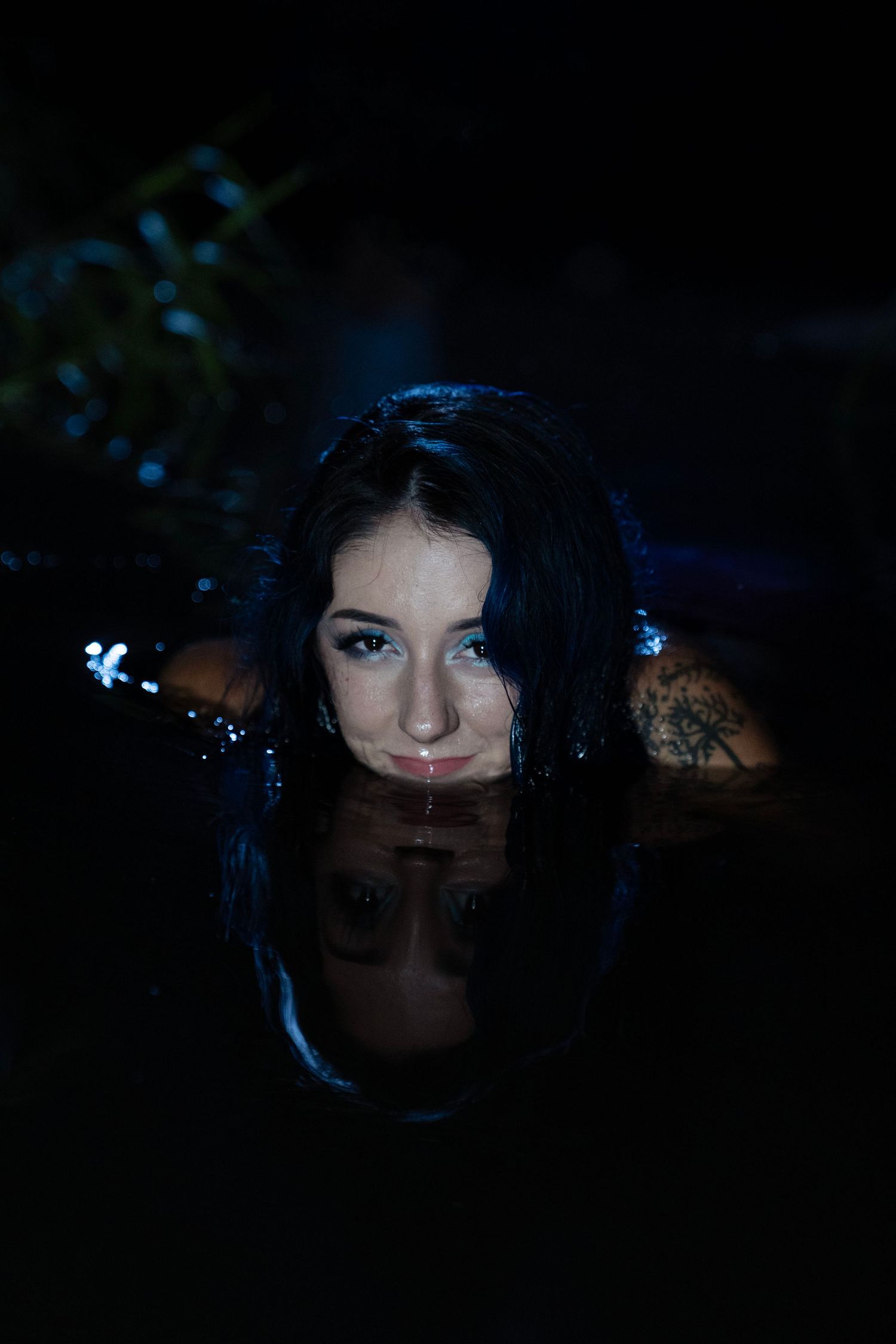Woman with dark wet hair and tattoo reflects in still dark water in a moody night portrait.