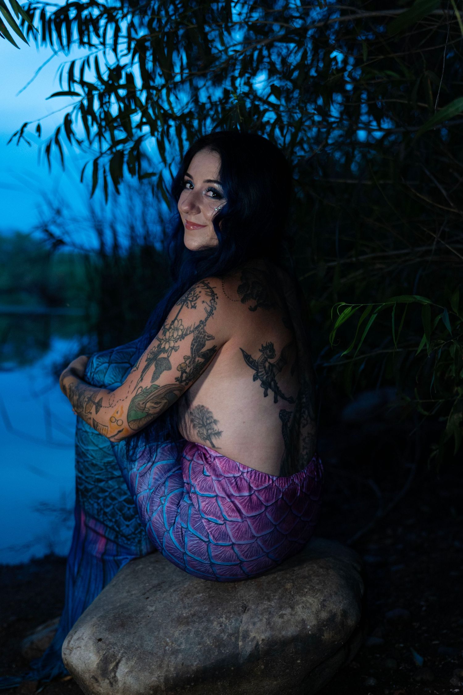 Tattooed woman in mermaid costume sitting on rocks by water at dusk.