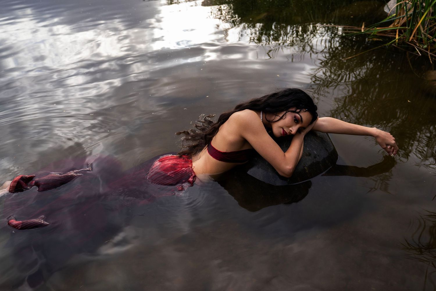 A woman with long dark hair lies on a rock in shallow water, wearing a red mermaid tail costume in a serene river setting.