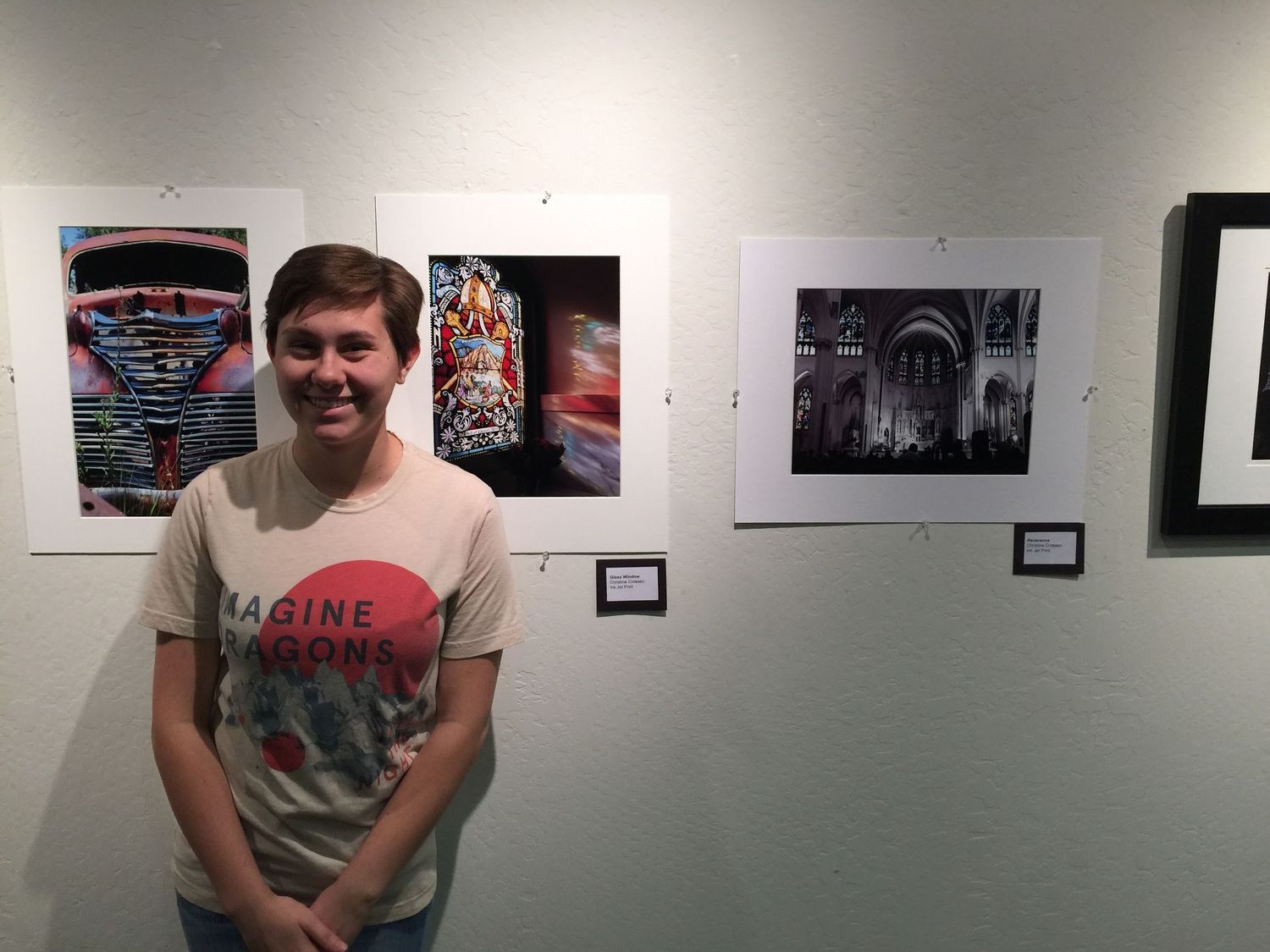 A smiling young person in a graphic t-shirt stands beside framed photographs displayed on a white gallery wall at an art exhibition.