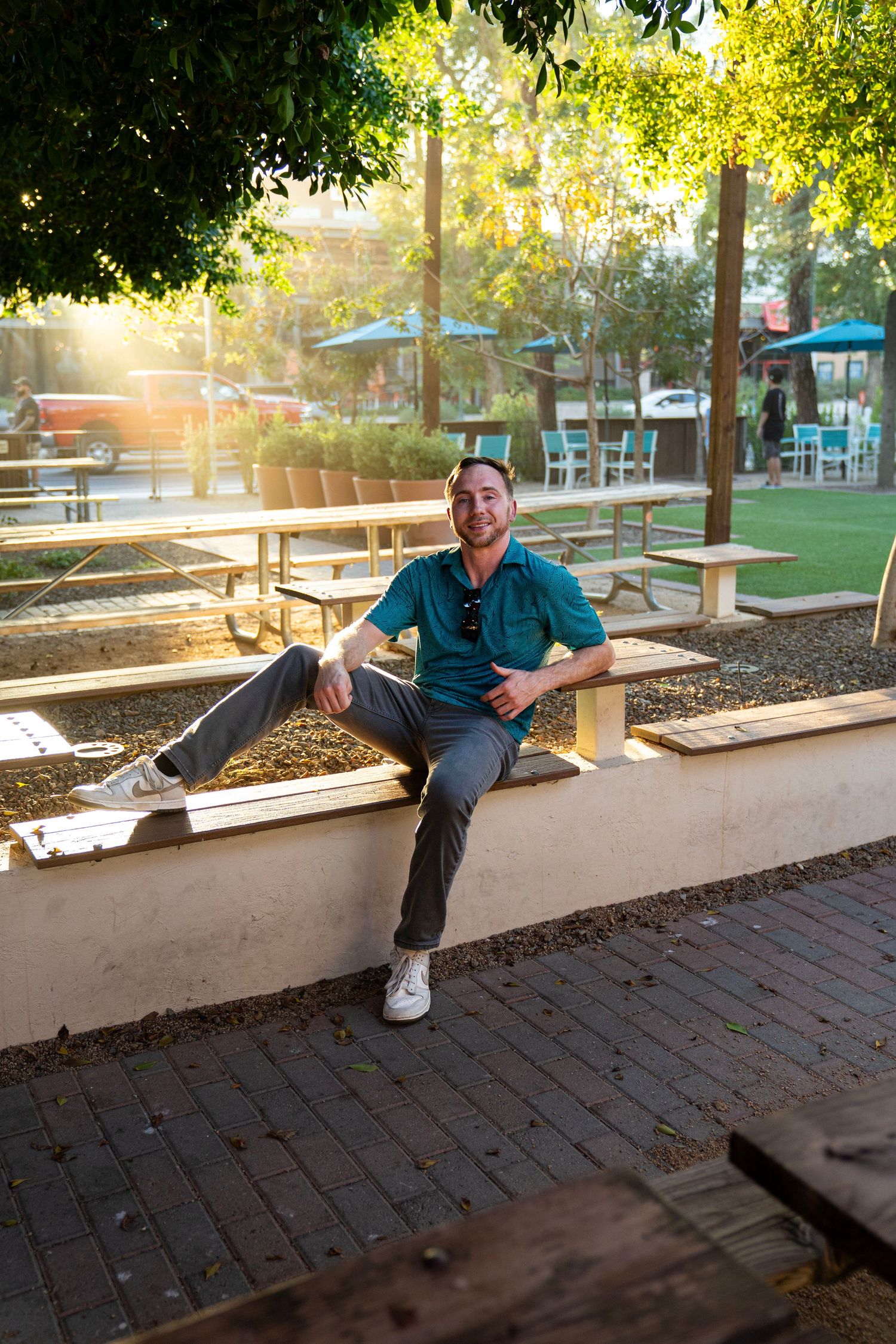 Man in teal polo shirt sitting on outdoor wall, smiling in sunlit patio area with picnic tables and trees.
