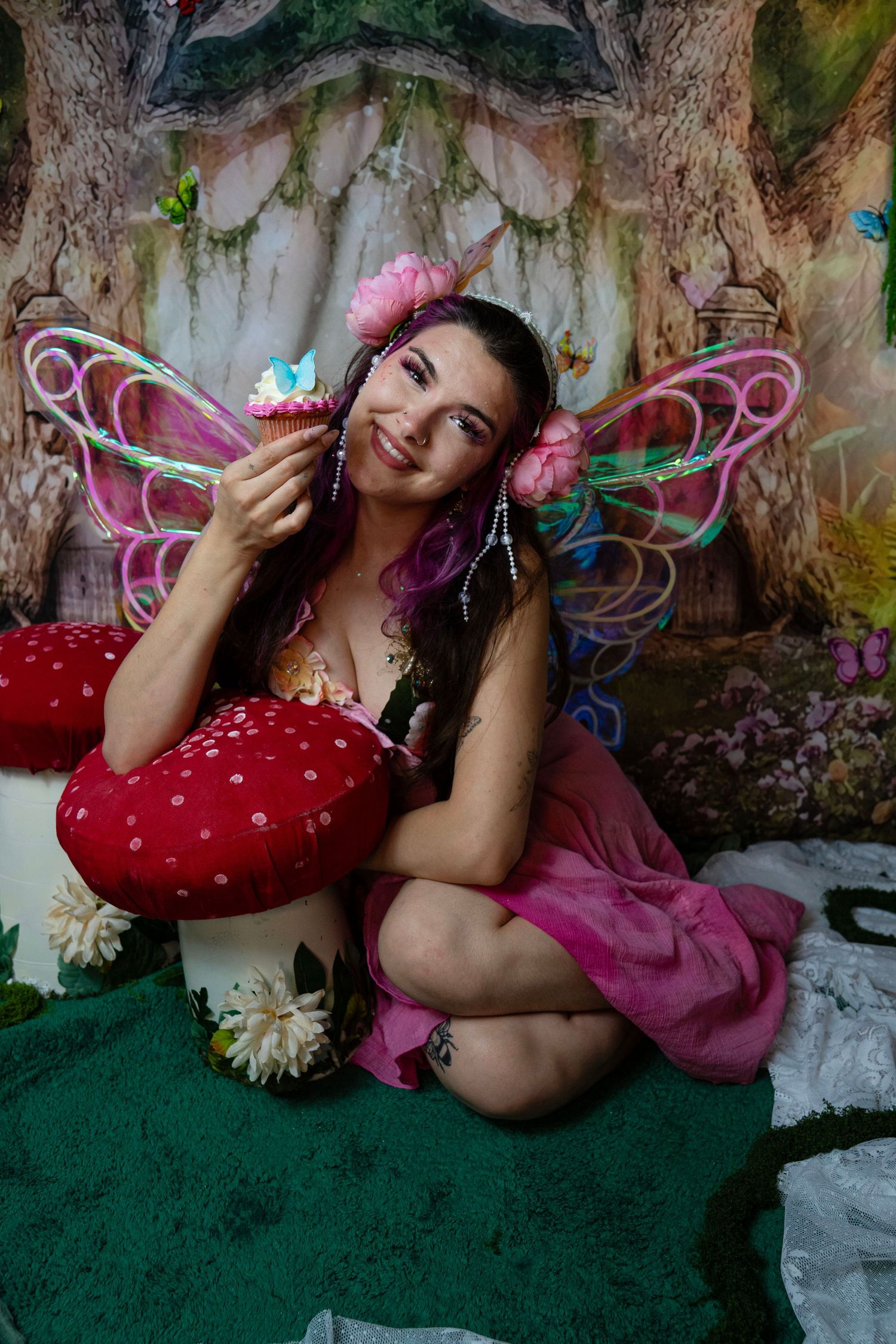 Smiling woman in pink fairy costume with butterfly wings holds a cupcake while sitting in a magical forest backdrop. Fantasy Photoshoot