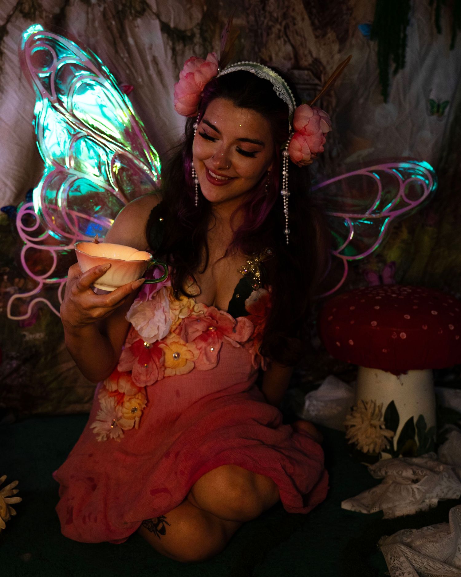 Smiling fairy costume model holds glowing candle cup beside red mushroom in dimly lit enchanted forest backdrop. Fantasy Photoshoot