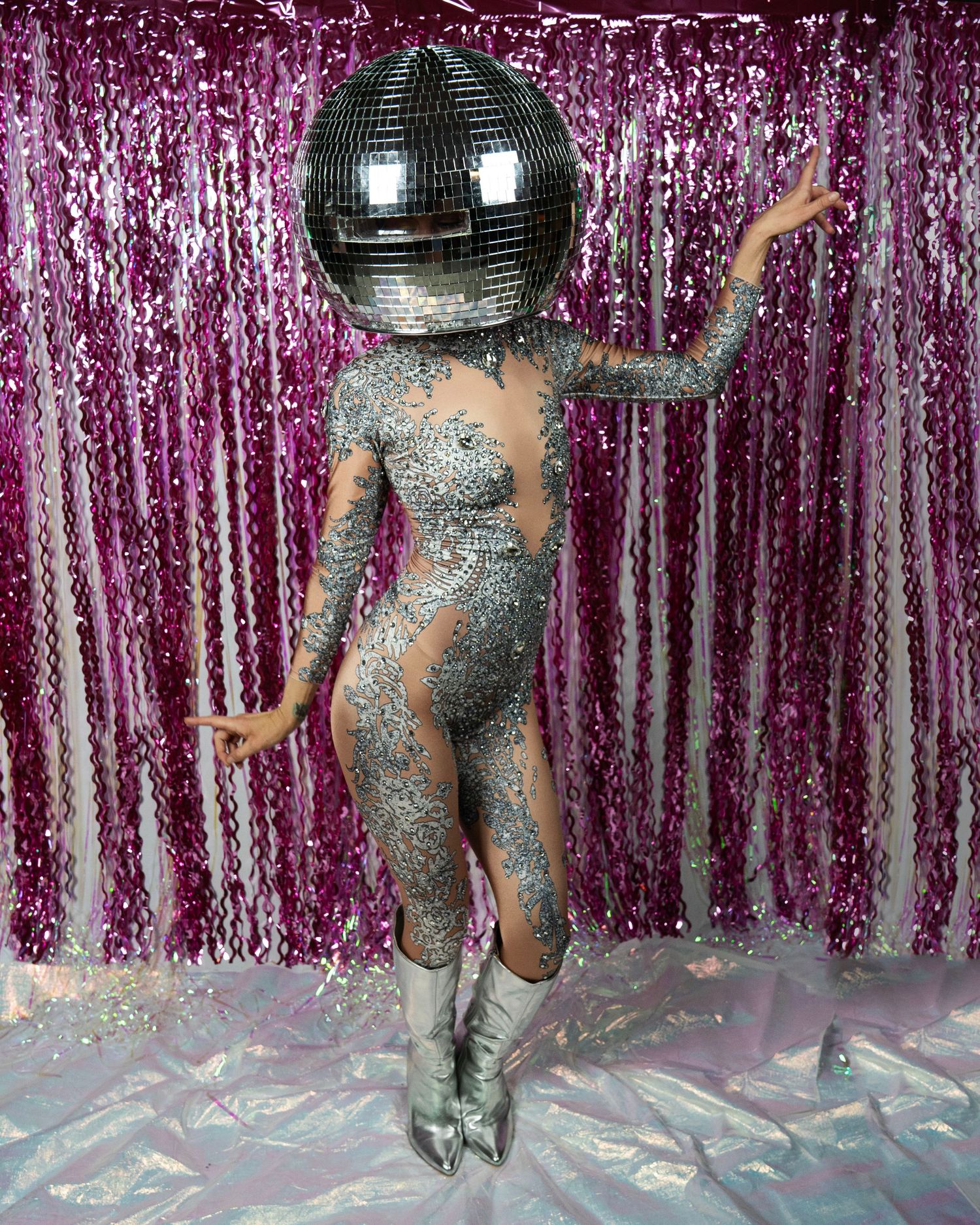 Dancing model in silver rhinestone bodysuit with disco ball head and silver boots on pink backdrop.