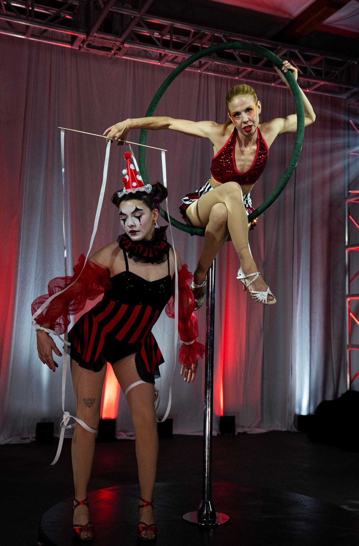 Circus clown and red-sequined aerialist perform coordinated pole hoop act on white-draped stage. Circus Photoshoot